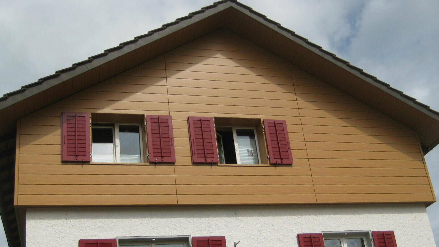 Hišna fasada v videzu lesa z vodoravno položenimi ploščami PREFA Siding, okna z rdečimi polkni
