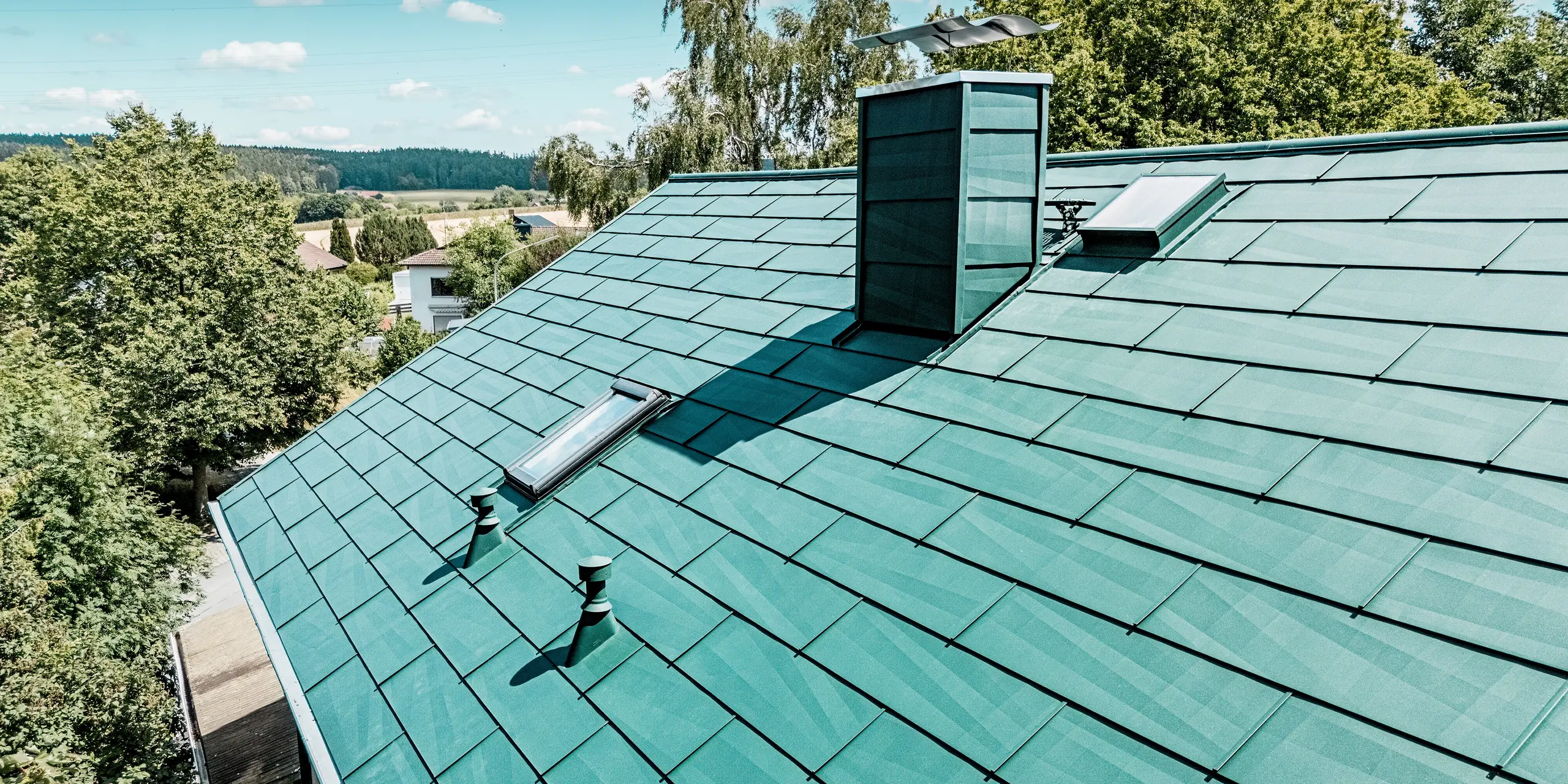 Modernisiertes Dach eines Einfamilienhauses in Schwarzenbach-Martinlamitz mit dem langlebigen PREFA Dachpaneel FX.12 in P.10 Moosgrün. Das detailreiche Bild zeigt das markante Aluminiumdach mit klaren Linien, strukturierter Oberfläche und integrierten Dachfenstern sowie einem formschön verkleideten Kamin. Die reflektierenden Lichtspiele auf den Paneelen unterstreichen das moderne Design. Dank der widerstandsfähigen Aluminiumoberfläche bietet das Dach optimalen Schutz vor Witterung, ist wartungsfrei und überzeugt durch eine stilvolle, zeitlose Ästhetik.