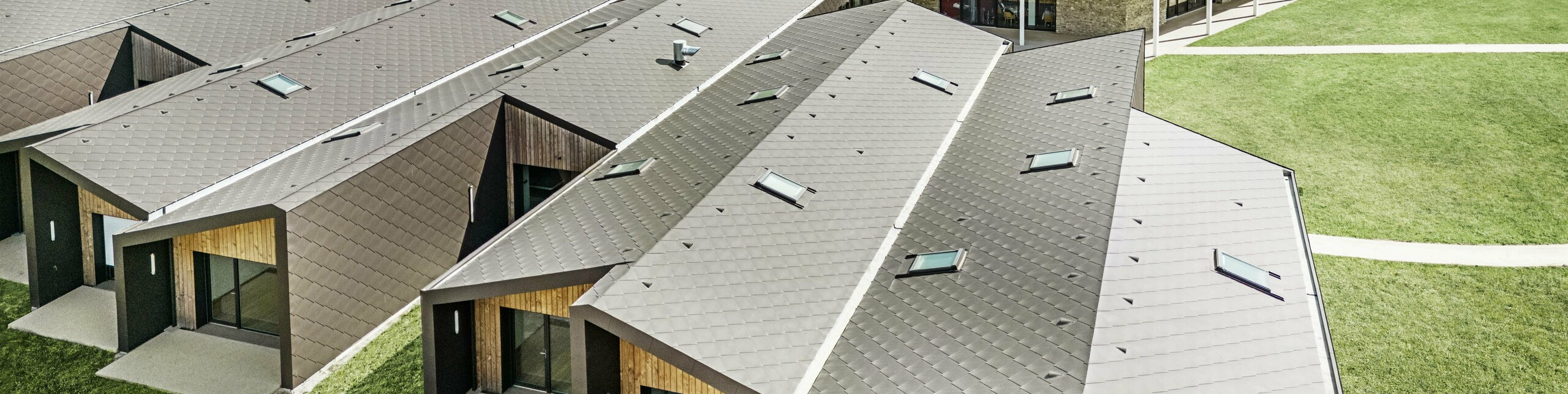 Slika prikazuje pogled iz zraka na sodoben stavbni kompleks, obložen s PREFA rombi 44 × 44 v barvi testa di moro. Za strešno površino so značilne geometrijske oblike v kombinaciji z velikimi steklenimi površinami in lesenimi elementi. V ozadju je vidna stara cerkev, ki tvori harmonično nasprotje sodobni arhitekturi. Zelena trata in okoliška narava poudarjata odprto in trajnostno zasnovo celotnega kompleksa.