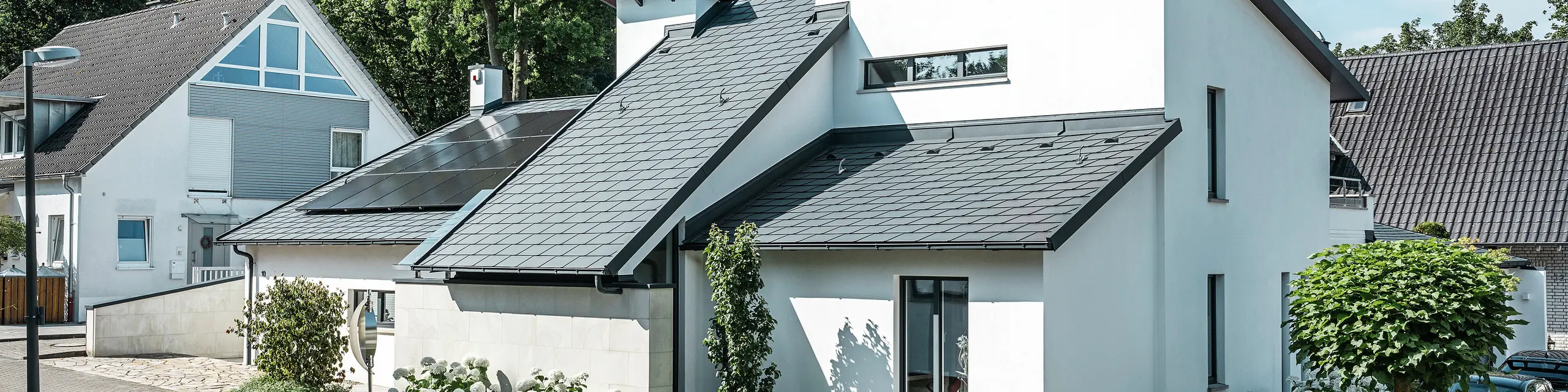 Moderna enodružinska hiša z belo fasado in temnim PREFA poševnim strešnim kritjem iz aluminija. Na strehi so vgrajeni fotovoltaični moduli in aluminijasti odvodnjavanje strehe v antracitni barvi. Stavba se nahaja v urejeni stanovanjski soseski z zasajenimi vrtovi, tlakovanim dovozom in okoliškimi enodružinskimi hišami.