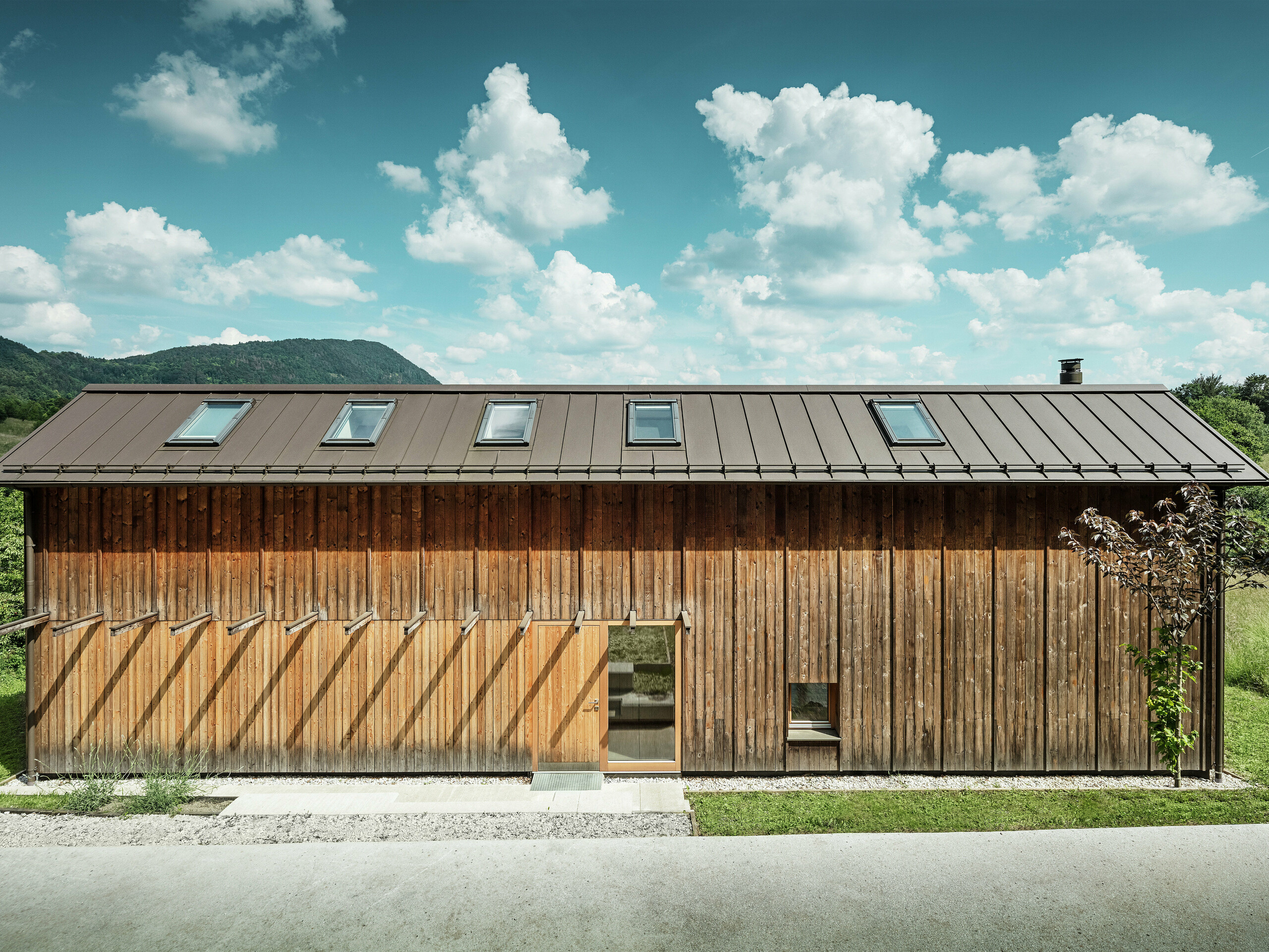 Ta sodobna hiša na pobočju navdušuje z minimalistično leseno fasado in vzdržljivo aluminijasto streho PREFALZ v rjavi barvi P.10. Strešna kritina s stoječimi zgibi navdušuje z visoko odpornostjo proti vremenskim vplivom in trajnostjo ter harmonično povezanostjo z naravnim okoljem. Vgrajena strešna okna in jasne linije poudarjajo brezčasno arhitekturo. Na sliki je prikazan vhodni del majhne samostojne hiše.