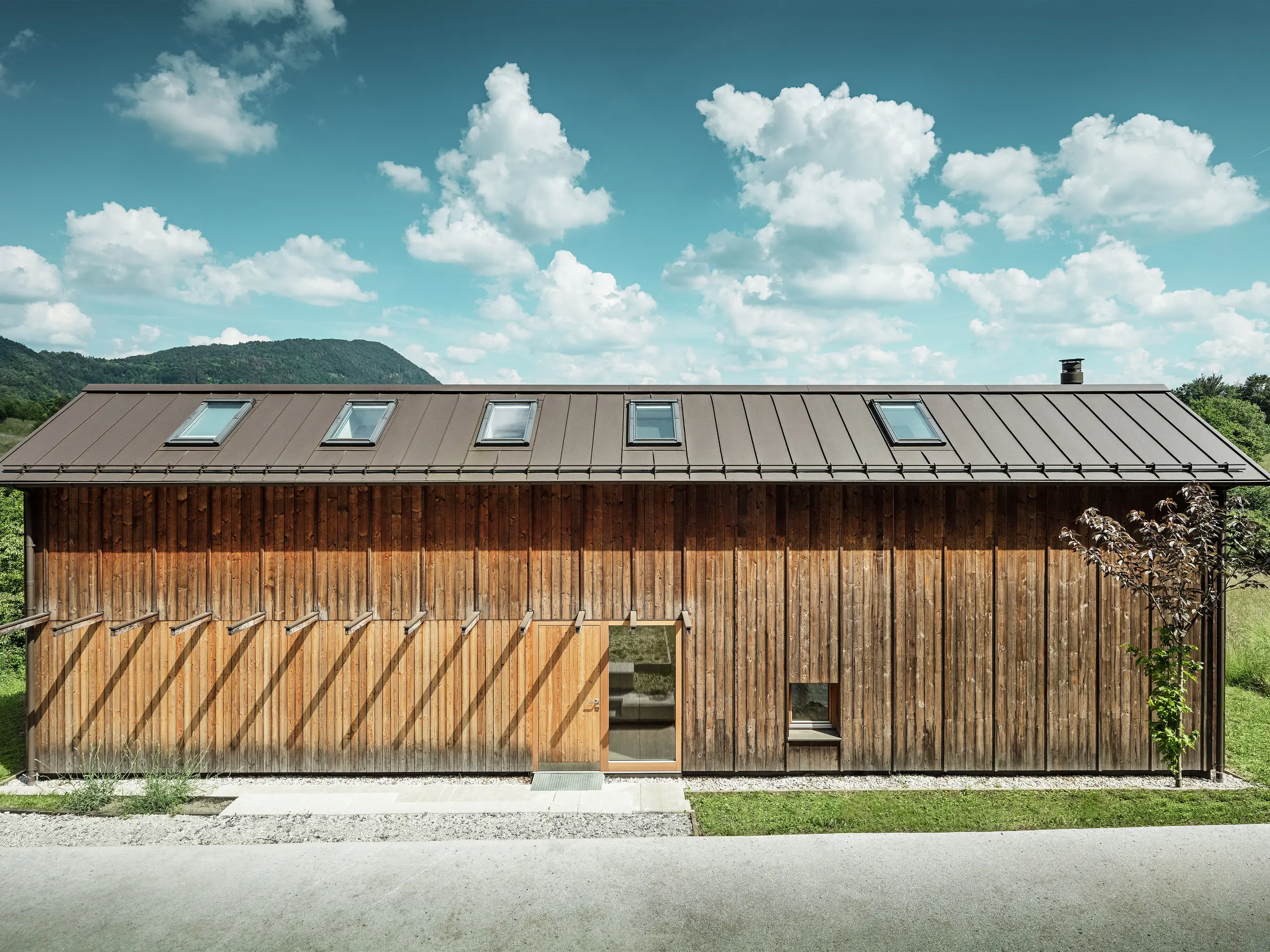 Ta sodobna hiša na pobočju navdušuje z minimalistično leseno fasado in vzdržljivo aluminijasto streho PREFALZ v rjavi barvi P.10. Strešna kritina s stoječimi zgibi navdušuje z visoko odpornostjo proti vremenskim vplivom in trajnostjo ter harmonično povezanostjo z naravnim okoljem. Vgrajena strešna okna in jasne linije poudarjajo brezčasno arhitekturo. Na sliki je prikazan vhodni del majhne samostojne hiše.