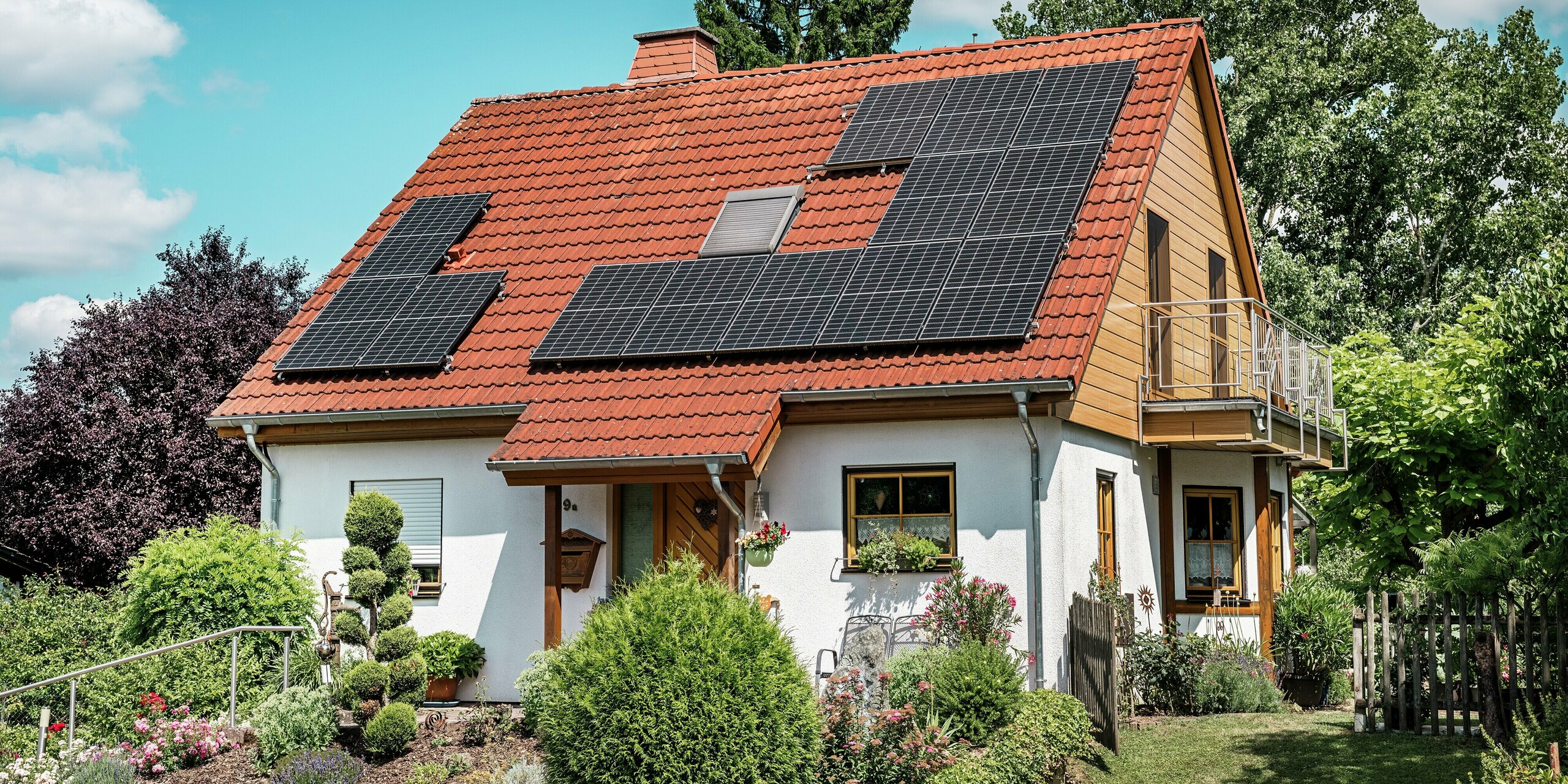 Das Bild zeigt ein charmantes Einfamilienhaus in Blieskastel, dessen frisch sanierter Giebel mit PREFA Sidings in Eiche Natur sofort ins Auge fällt. Die warme Holzoptik des Aluminiums harmoniert eindrucksvoll mit dem roten Ziegeldach und den großflächigen PV-Modulen. Umgeben von einem liebevoll gepflegten Garten entsteht ein lebendiges, sonnendurchflutetes Gesamtbild. Die witterungsbeständigen und farbstabilen PREFA Sidings bieten die natürliche Ästhetik von Holz – jedoch völlig wartungsfrei und dauerhaft formstabil. Ideal für langlebige Fassadensanierungen.
