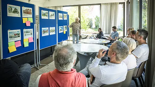 PREFARENZEN 2027: strokovni dialog v svetli, moderni seminarni dvorani. Moderator Matthias Finkentey stoji pred modrimi oglasnimi tablami, na katerih so predstavljeni arhitekturni projekti z uporabo PREFA aluminijastih rešitev. Žirija sedi pozorno za okroglimi mizami, razpravlja in si zapisuje vtise. Velika okna prepuščajo dnevno svetlobo, medtem ko je skozi njih viden zelen vrt. Prizor prikazuje povezavo med ustvarjalno arhitekturno razpravo, funkcionalnim načrtovanjem in ocenjevanjem oblikovanja. PREFA aluminijasti izdelki predstavljajo trajne, estetsko dovršene strehe in fasade, ki v arhitekturnih projektih utelešajo inovativnost, kakovost in navdih.
