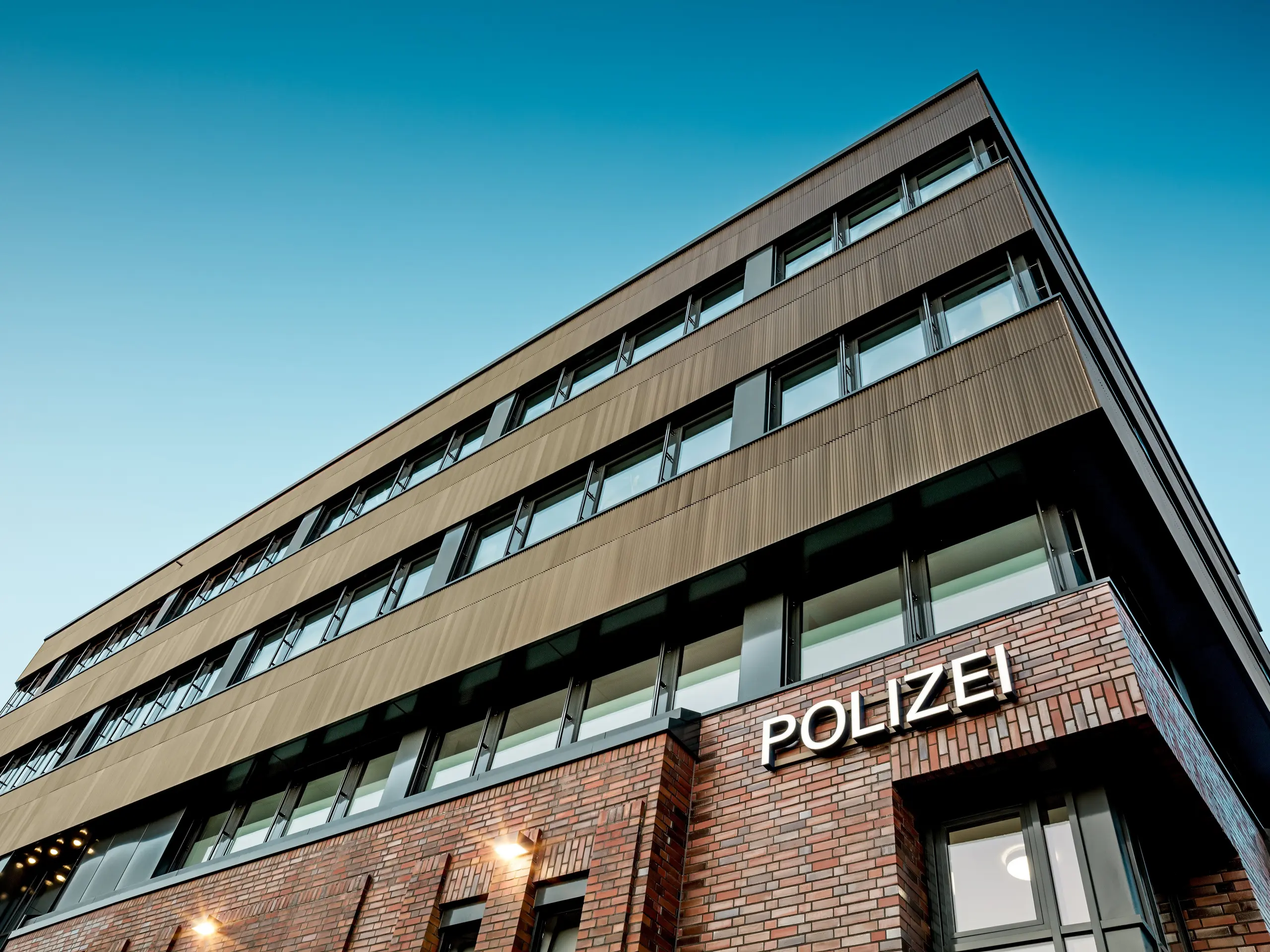 Arhitekturni pogled od spodaj na policijsko postajo 43 v Hamburgu: nad obstoječo zgradbo iz opeke se dviga natančno strukturirana nova stavba s PREFA aluminijasto fasado iz ekstrudiranih zobčastih profilov v elegantni barvi srednje bronaste. Fina rebrasta površina pritegne pogled in ustvarja fascinantne svetlobne odseve vzdolž jasnih, vodoravnih pasov oken. PREFA aluminij zagotavlja najvišjo odpornost proti koroziji, dolgo življenjsko dobo in minimalno vzdrževanje – trajnostna rešitev z izrazitim dizajnom za moderno in funkcionalno fasadno arhitekturo.