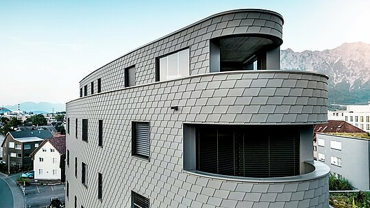 Podroben stranski pogled na hišo Bernerhaus v Buchsu z mehko zaobljenimi koti in neprekinjenimi balkonskimi trakovi. Fasada iz PREFA fasadnih skodel v barvi P.10 svetlo siva se kot natančno obdelana aluminijasta koža ovije okoli trikotne kubature in ustvarja živahen, luskast vzorec. Enotna površina poudarja tekoče zaobljenosti ter igro svetlobe in sence. Zahvaljujoč lahkemu, vremensko odpornemu in trajnemu PREFA aluminiju nastane fasada, ki ne potrebuje veliko vzdrževanja in ima izrazit videz na daljavo pred alpskim ozadjem.