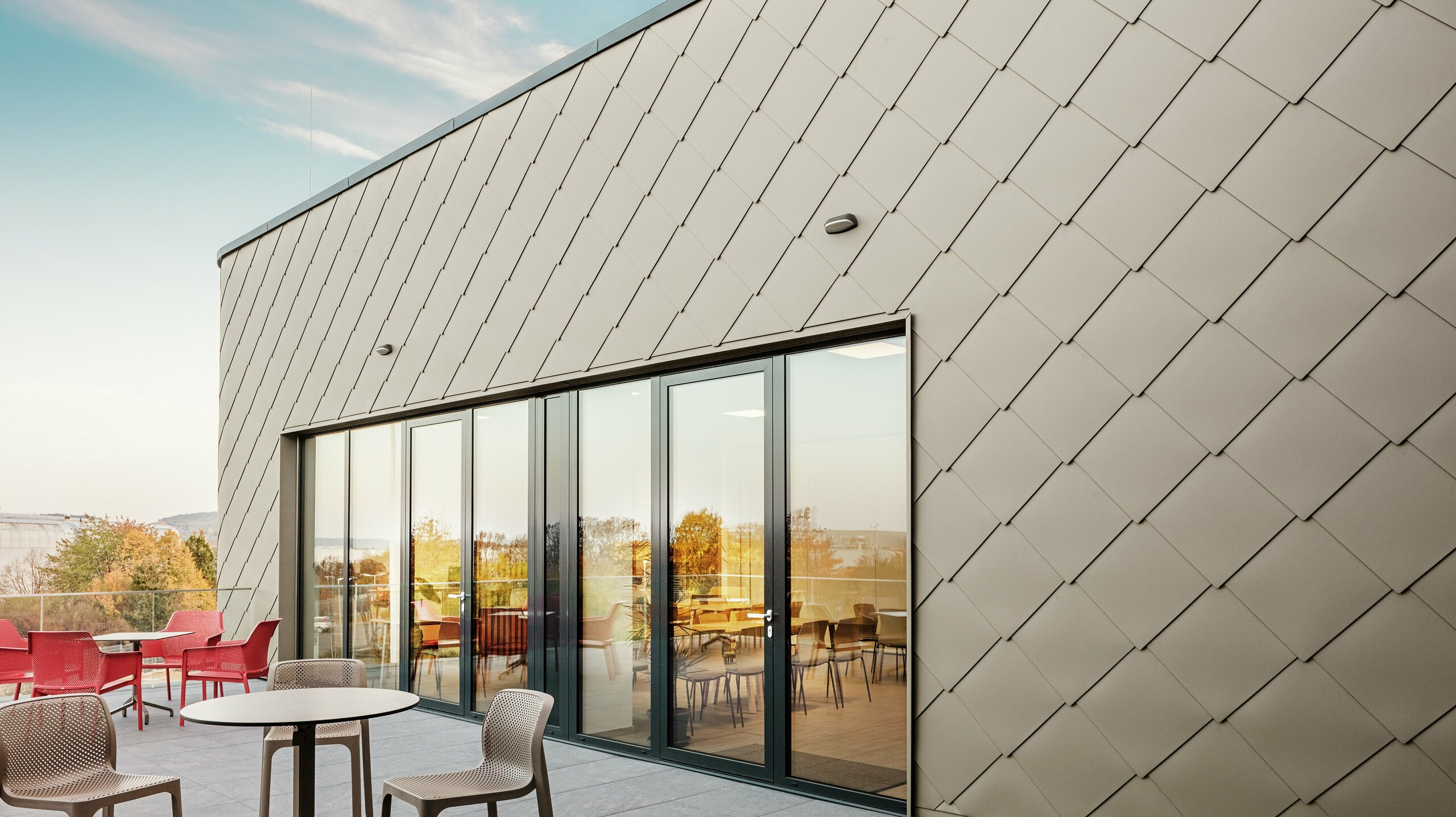 Fasada nove PREFA lokacije v Törökbálintu navdušuje z elegantnimi aluminijastimi rombi PREFA 44 × 44 v bronasti barvi. Natančno izvedena aluminijasta fasada ustvarja sodoben in dinamičen videz ter hkrati nudi vrhunsko zaščito pred vremenskimi vplivi. Prostorna steklena fronta povezuje notranjost z zunanjostjo in ustvarja svetlo ter prijetno vzdušje na veliki terasi.