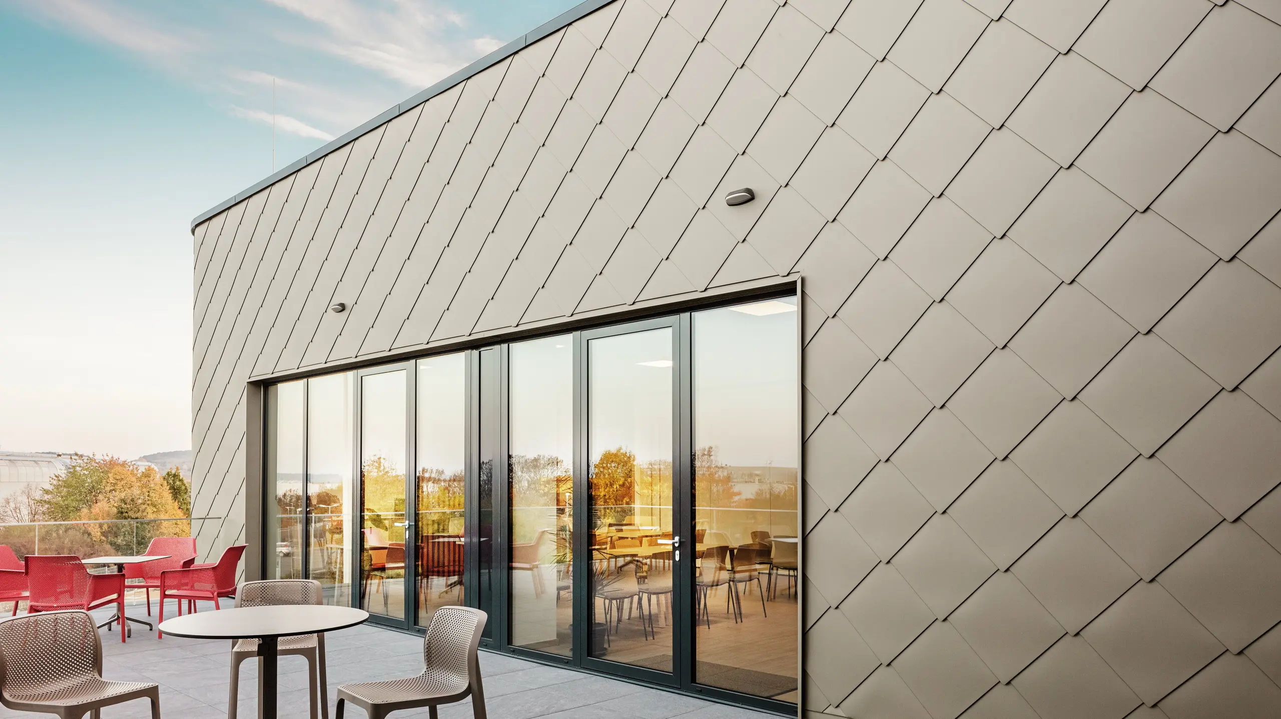 Fasada nove PREFA lokacije v Törökbálintu navdušuje z elegantnimi aluminijastimi rombi PREFA 44 × 44 v bronasti barvi. Natančno izvedena aluminijasta fasada ustvarja sodoben in dinamičen videz ter hkrati nudi vrhunsko zaščito pred vremenskimi vplivi. Prostorna steklena fronta povezuje notranjost z zunanjostjo in ustvarja svetlo ter prijetno vzdušje na veliki terasi.