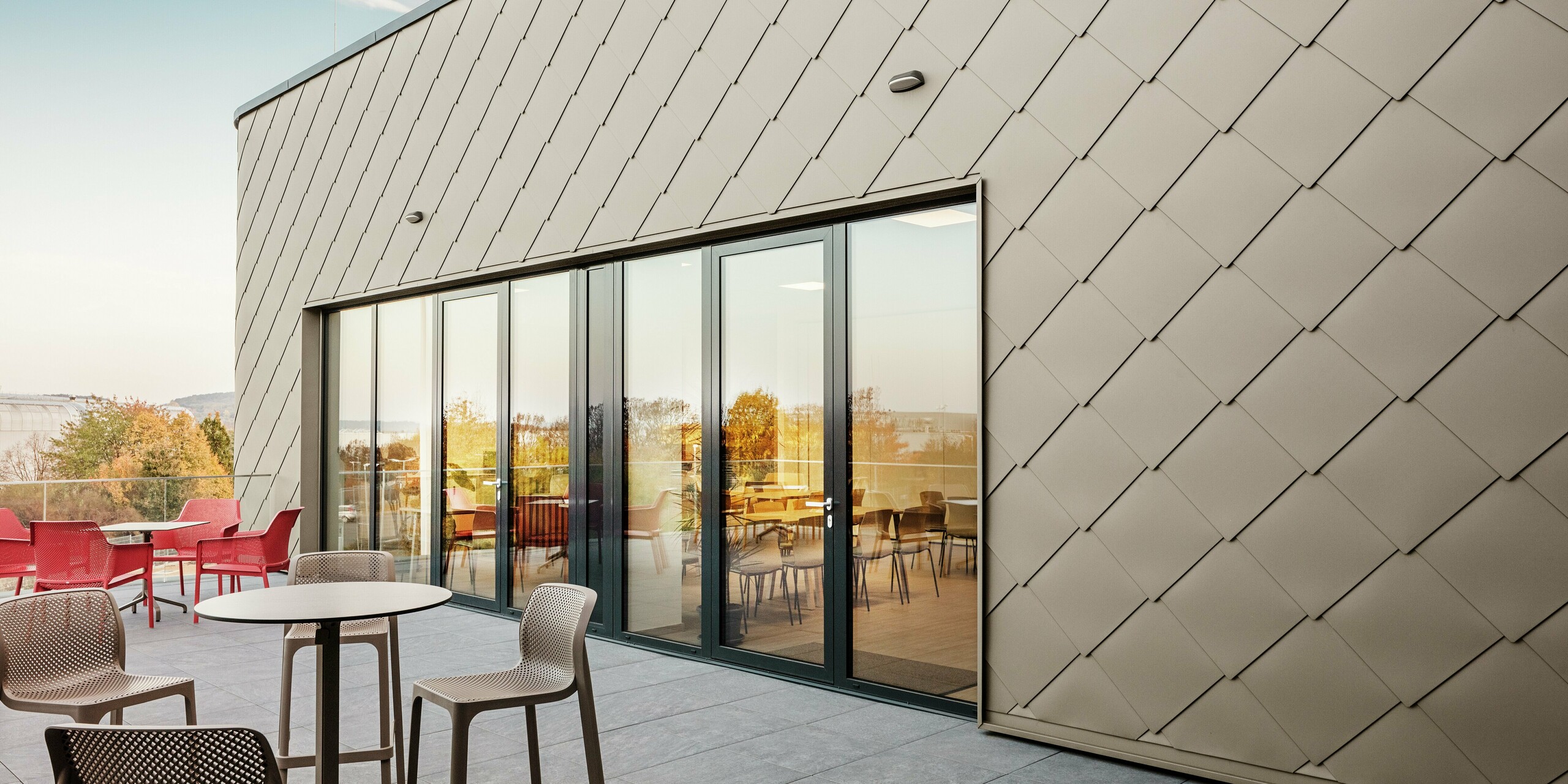 Fasada nove PREFA lokacije v Törökbálintu navdušuje z elegantnimi aluminijastimi rombi PREFA 44 × 44 v bronasti barvi. Natančno izvedena aluminijasta fasada ustvarja sodoben in dinamičen videz ter hkrati nudi vrhunsko zaščito pred vremenskimi vplivi. Prostorna steklena fronta povezuje notranjost z zunanjostjo in ustvarja svetlo ter prijetno vzdušje na veliki terasi.