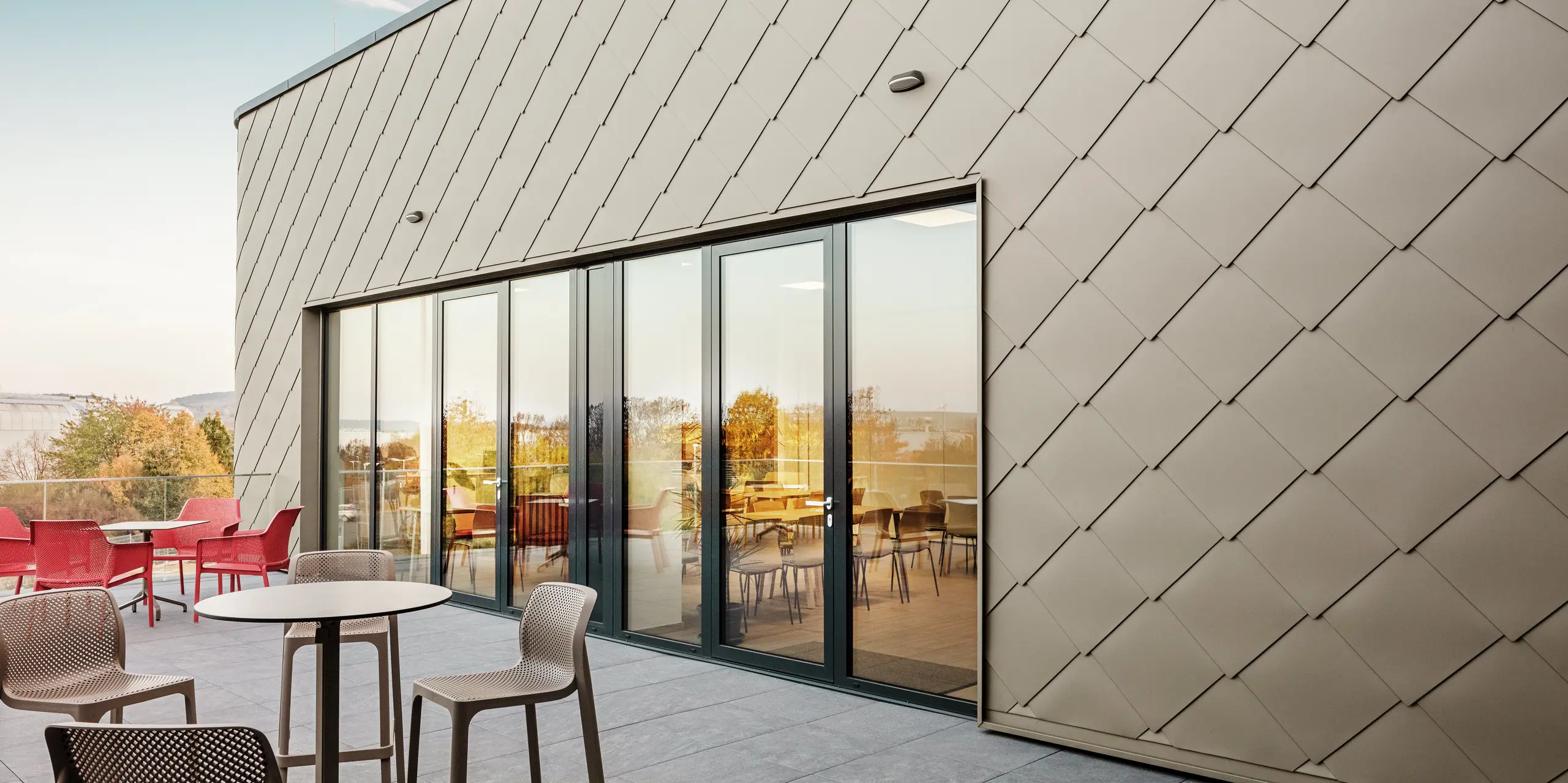 Fasada nove PREFA lokacije v Törökbálintu navdušuje z elegantnimi aluminijastimi rombi PREFA 44 × 44 v bronasti barvi. Natančno izvedena aluminijasta fasada ustvarja sodoben in dinamičen videz ter hkrati nudi vrhunsko zaščito pred vremenskimi vplivi. Prostorna steklena fronta povezuje notranjost z zunanjostjo in ustvarja svetlo ter prijetno vzdušje na veliki terasi.