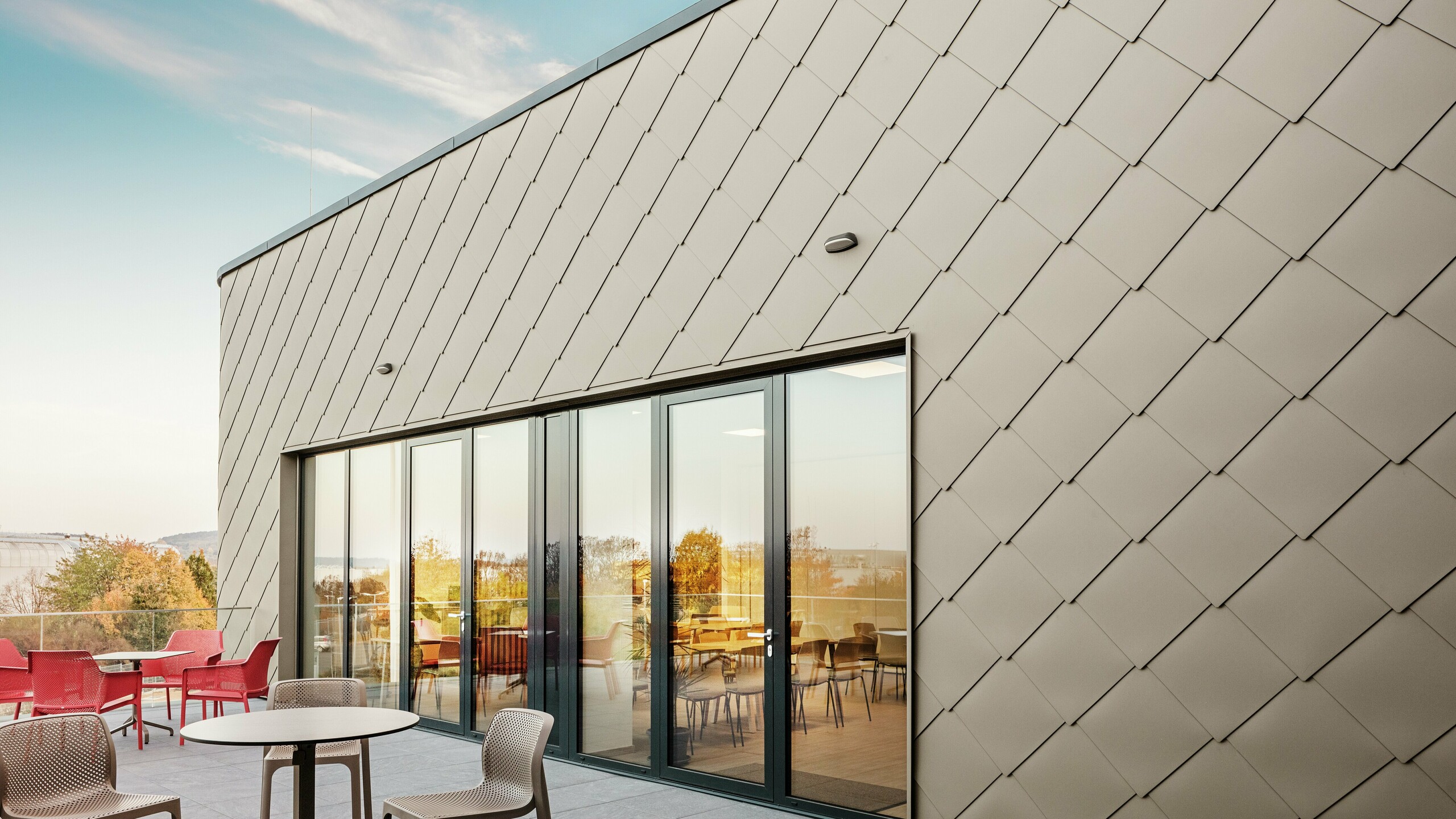 Fasada nove PREFA lokacije v Törökbálintu navdušuje z elegantnimi aluminijastimi rombi PREFA 44 × 44 v bronasti barvi. Natančno izvedena aluminijasta fasada ustvarja sodoben in dinamičen videz ter hkrati nudi vrhunsko zaščito pred vremenskimi vplivi. Prostorna steklena fronta povezuje notranjost z zunanjostjo in ustvarja svetlo ter prijetno vzdušje na veliki terasi.