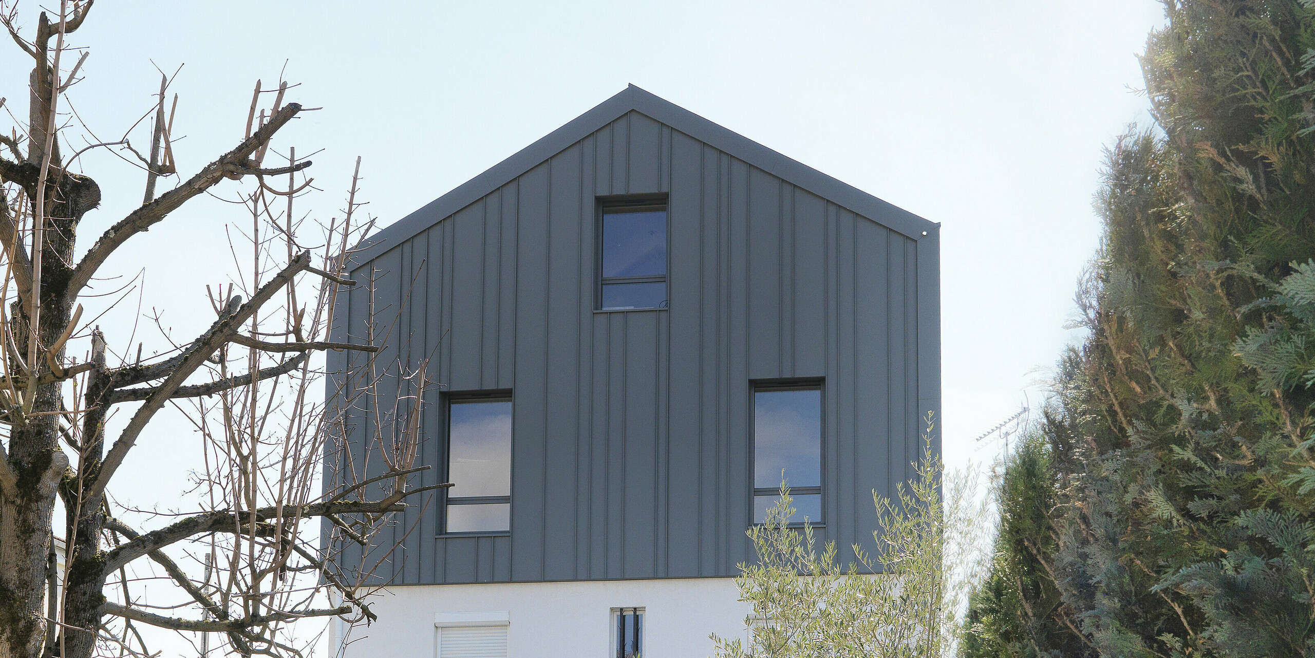 Pogled iz vrta na enodružinsko hišo v Houillesu v Franciji. Streha in fasada hiše sta prekriti s kombinacijo elementov PREFA Siding v P.10 beli barvi in trakovi PREFALZ v P.10 svetlo sivi barvi.