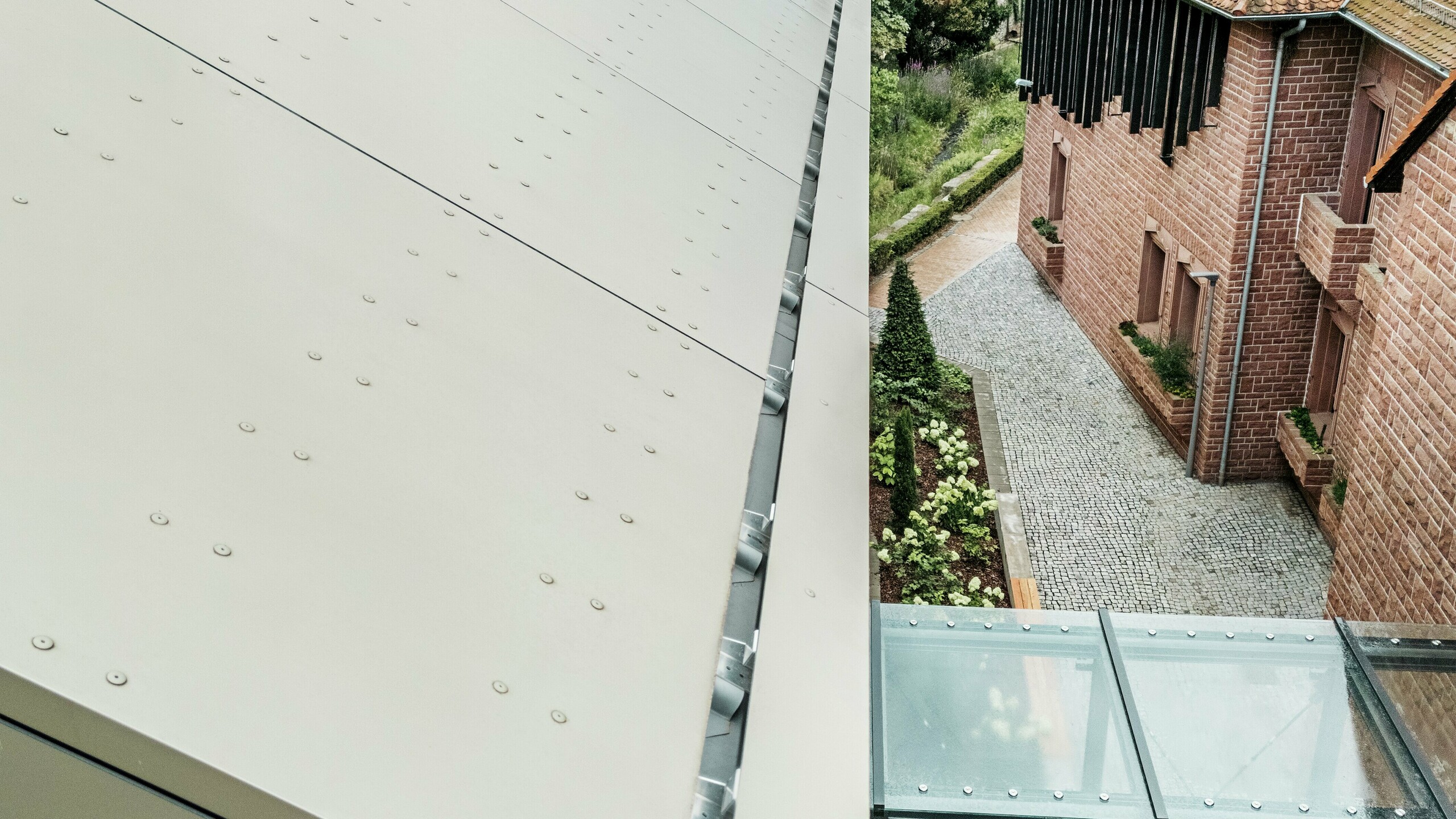 Podrobni posnetek strehe mestne hiše v Alzenau, ki je pokrita z aluminijastimi kompozitnimi ploščami PREFA v nežnem bronastem odtenku. Plošče so natančno položene in opremljene z vidnimi pritrdilnimi elementi, kar fasadi daje sodoben, industrijski videz. V ospredju je vidna steklena povezovalna galerija, ki mestno hišo povezuje s sosednjo stavbo.