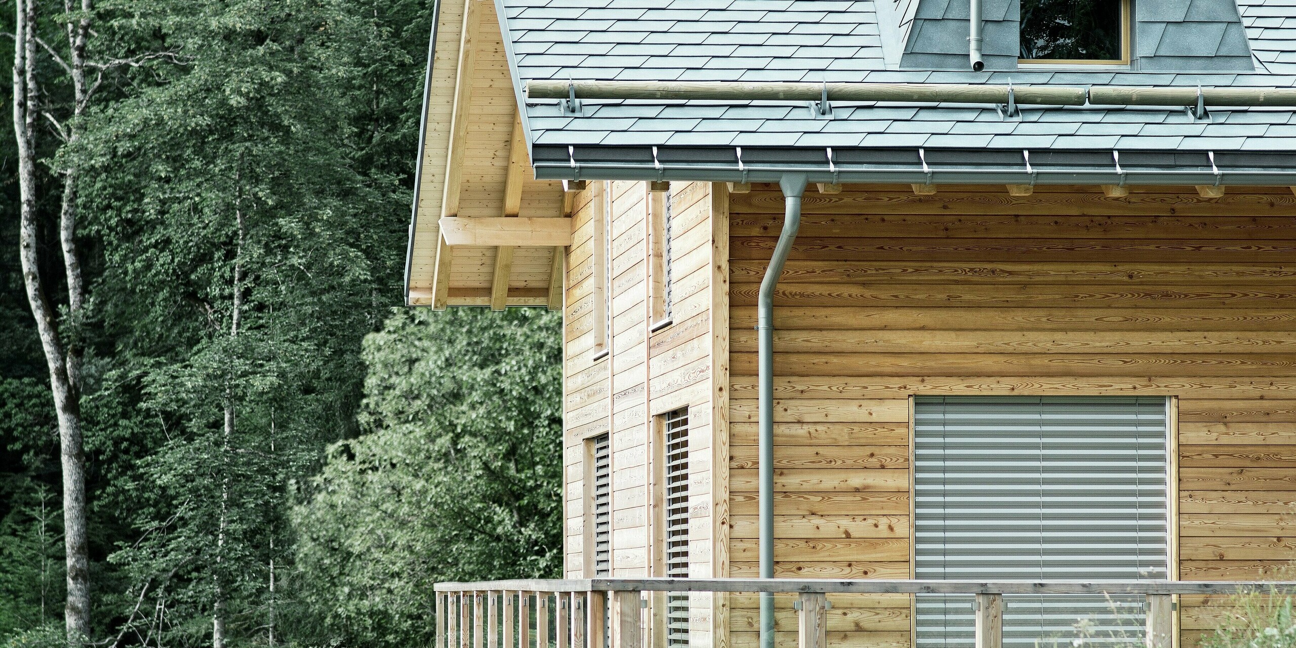 Seitenansicht einer Holz-Lodge am Genfersee mit PREFA Aluminium-Dachschindeln in Steingrau. Die Dachstruktur mit dem markanten Kamin und dem Schneefang aus Rundholz fügt sich harmonisch in die Umgebung ein. Das langlebige und ästhetische Blechdach bietet nicht nur Schutz vor den Elementen, sondern stellt auch eine stilvolle Ergänzung zur Holzfassade dar.