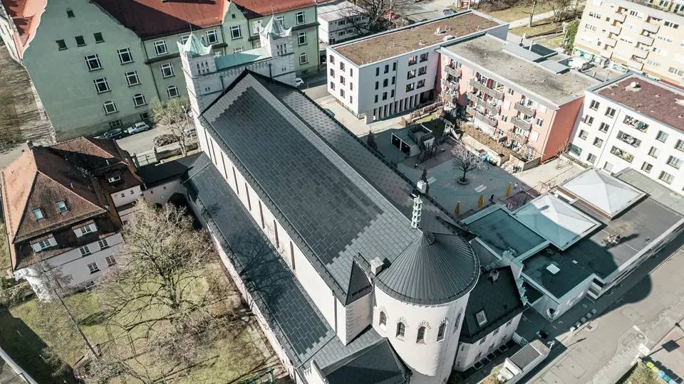 Pogled iz ptičje perspektive na zadnjo stran cerkve sv. Karla Borromeja v Nürnbergu. Črne solarne strešne plošče PREFA pokrivajo značilno, okroglo dvokapno streho nad glavnim delom cerkve ter streho stranskega objekta. Aluminijaste strešne plošče z vgrajenimi fotovoltaičnimi moduli se odlično vklapljajo v zgodovinski videz cerkve. Pogled na sodobno okolico s sosednjimi stanovanjskimi stavbami in zelenim dvoriščem ponazarja uspešno povezavo med varstvom kulturne dediščine, sodobno arhitekturo in trajnostnim pridobivanjem energije. Idealna rešitev za varstvo kulturne dediščine v urbanem okolju.