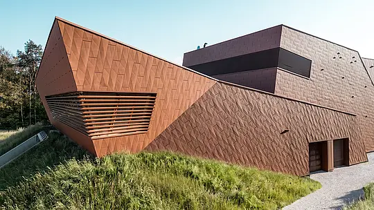 Izrazita črpalnica za pitno vodo WML v Lottumu z dinamično zloženo stavbo in izrazitim vrhom, ki pritegne pogled. Aluminijasta fasada PREFA Siding.X v topli rjavi barvi navdušuje s svojo faciranim, živahno strukturo ter natančnim prepletanjem svetlobe in sence. Vodoravne lamele poudarjajo globino arhitekture, hkrati pa se stavba harmonično vključuje v naravno okolje. Lahki, trajni in reciklirani PREFA aluminijasti paneli zagotavljajo najvišjo odpornost proti vremenskim vplivom ter trajnostno, vzdrževanju prijazno ovojnico stavbe.