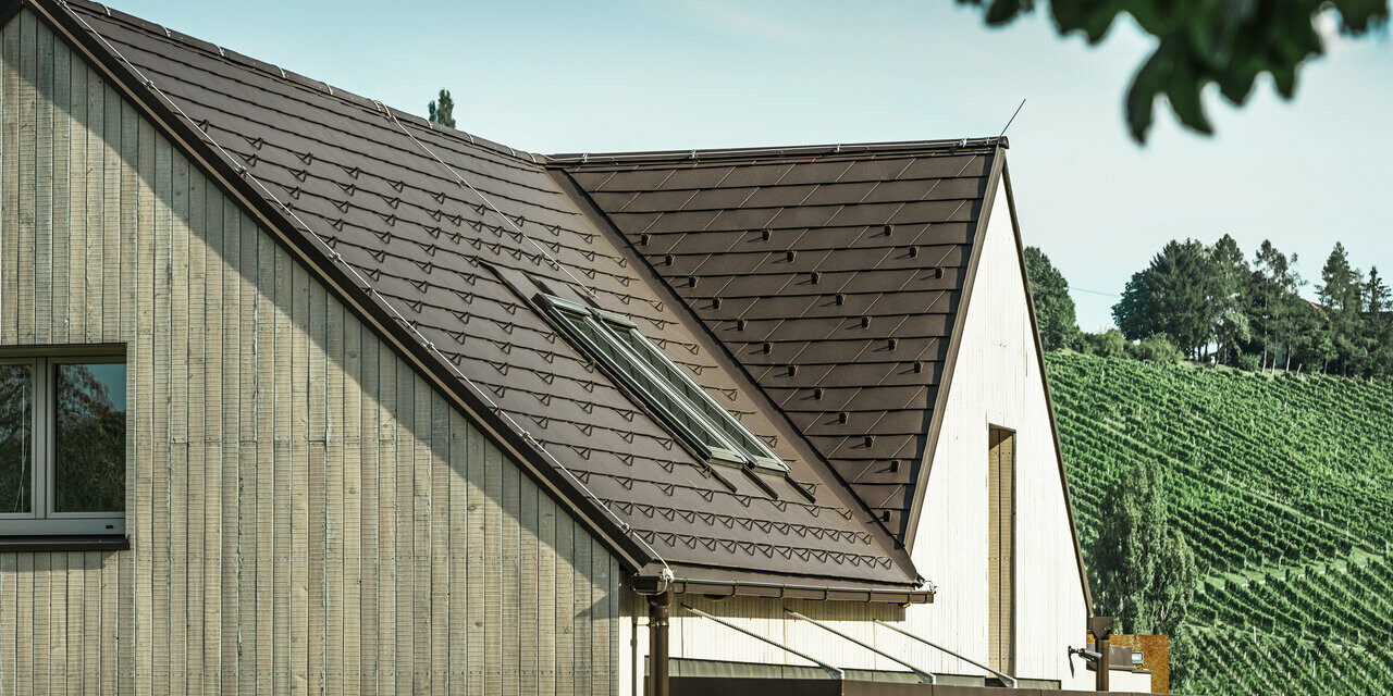 Enodružinska hiša z dvema dvokapnima strehama je prekrita s strešnimi skodlami PREFA v barvi testa di moro. Za odvodnjavanje strehe se uporablja pravokotni žleb PREFA. Fasada je obložena z lesom.