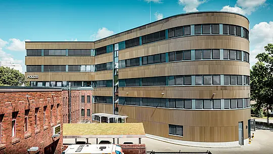 Dinamičen zunanji pogled na razširjeno policijsko postajo 43 v Hamburgu: ukrivljena nova stavba s približno 1.100 m² PREFA aluminijaste fasade iz eloksiranih ekstrudiranih zobčastih profilov v barvi srednje bronaste je prava paša za oči in ustvarja izrazit kontrast z obstoječo zgradbo iz opeke. Fino strukturirana površina živahno igra s svetlobo in senco ter fasadi daje globino in natančnost. Zahvaljujoč visoki odpornosti proti koroziji, dolgoživosti in lastnostim, ki zahtevajo le malo vzdrževanja, PREFA aluminijaste rešitve zagotavljajo trajnostno, funkcionalno in arhitekturno zahtevno kakovost gradnje.