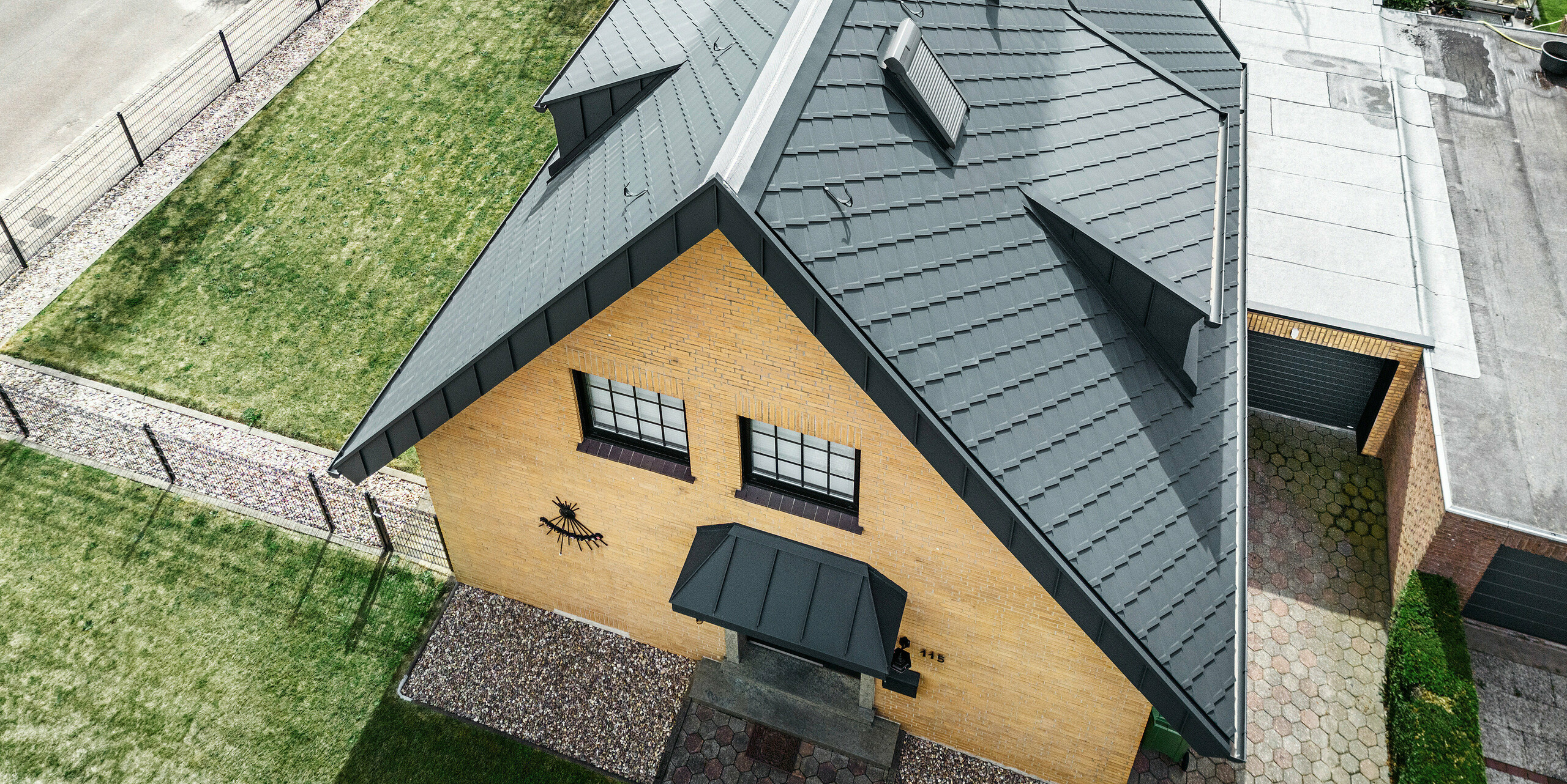 Pogled iz ptičje perspektive na enodružinsko hišo s PREFA streho antracitne barve s strešnimi rombi 29×29 in komplementarnim sistemom odvodnjavanja strehe. Hiša predstavlja fasado iz rumene opeke in dve mansardni okni, ki skupaj s streho ustvarjata skladen estetski kontrast. Celoten pogled kaže na skrbno razporeditev strešnih elementov in strukturirano eleganco zasnove strehe.