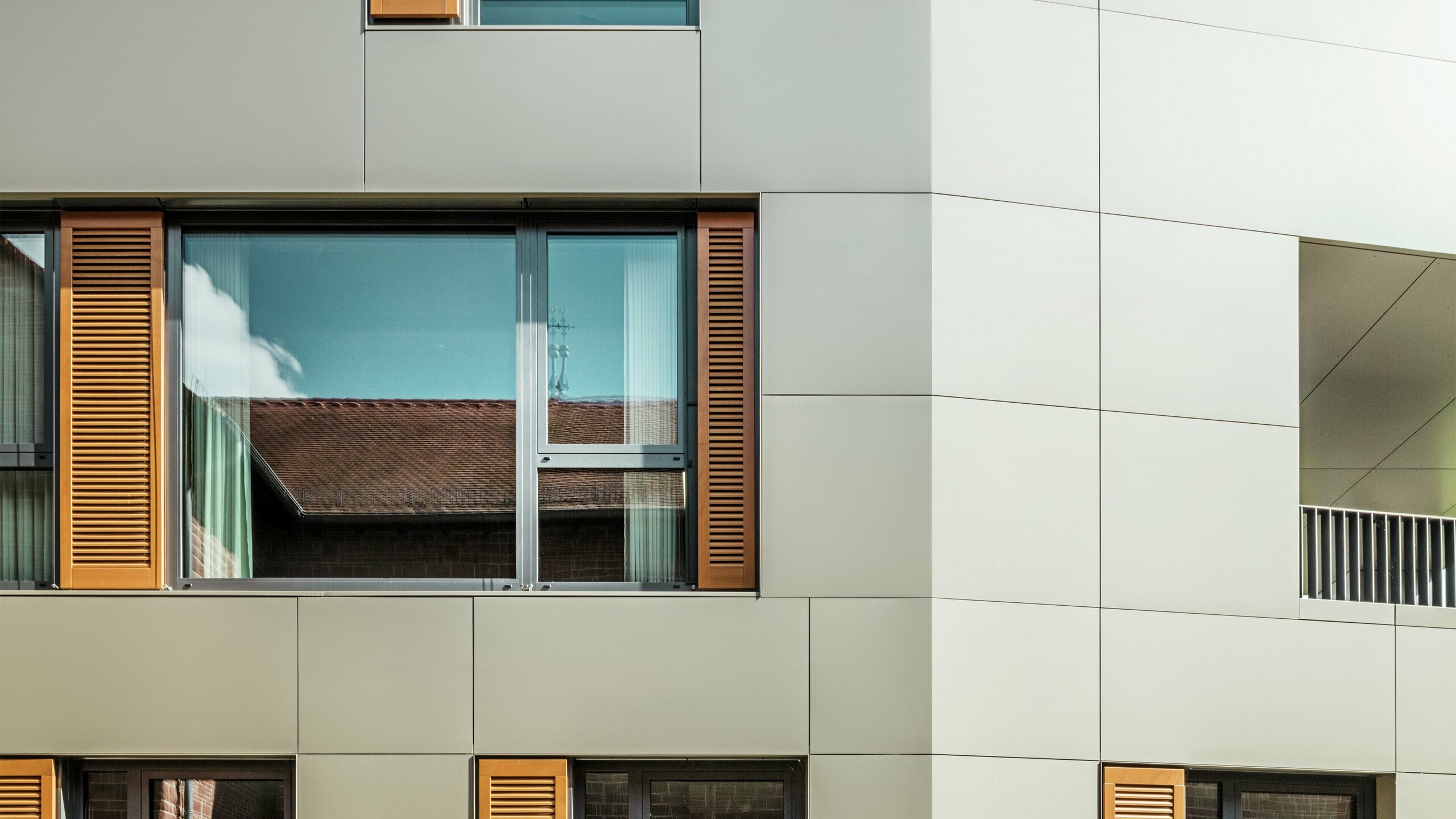 Podrobni posnetek fasade mestne hiše v Alzenauu v Nemčiji z aluminijastimi kompozitnimi ploščami PREFA v bronasti barvi. Velika okna obdajajo elegantne lesene žaluzije v topli rjavi barvi, ki fasadi dajejo privlačno teksturo. V ospredju je vidna urejena cvetlična greda, ki z zelenimi poudarki dopolnjuje urbano obliko stavbe. Jasno nebo in čiste linije arhitekture poudarjajo sodoben značaj stavbe.