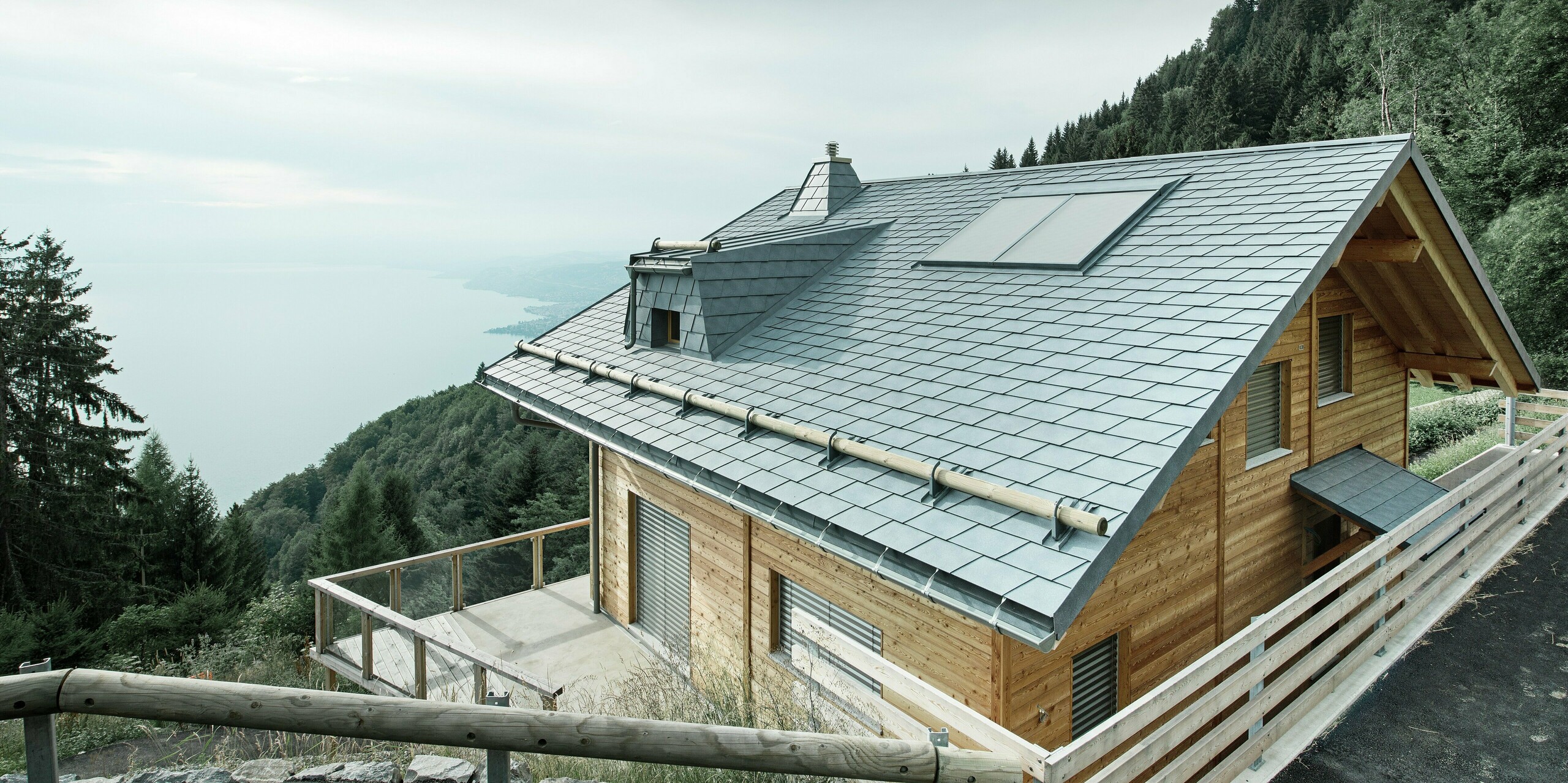 Dachansicht einer Holz-Lodge am Genfersee mit PREFA Dachschindeln in P.10 Steingrau. Die präzise verarbeitete Dachgaube und der Kamin in Trapezform setzen markante Akzente, während das robuste Dach Wind und Wetter trotzt. Das elegante Design aus Aluminium in Kombination mit Holz sorgt für eine perfekte Harmonie zwischen Funktion und Ästhetik. Im Hintergrund ist der in mystischen Nebel gehüllte Genfersee zu erkennen.
