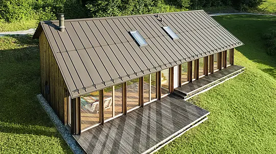 Sodobna lesena hiša z veliko stekleno fasado in trpežno aluminijasto streho PREFALZ v rjavi barvi P.10 - idealna za pobočja, kot je Vinje. Visokokakovostna strešna kritina zagotavlja največjo odpornost proti vremenskim vplivom, dolgo življenjsko dobo in eleganten videz. Natančna namestitev stoječih zgibov in vgrajena strešna okna ustvarjajo trajnostni bivalni koncept, z veliko svetlobe. Pogled s ptičje perspektive prikazuje natančno izdelavo aluminijastih stoječih zgibov.
