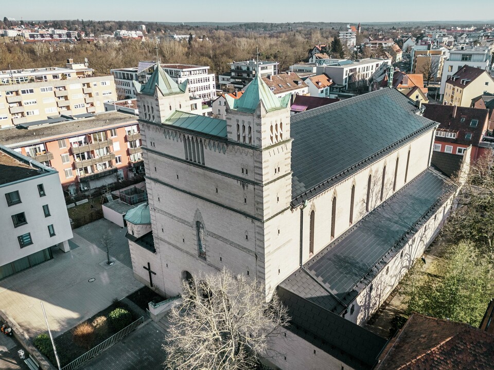 Vorderseite der St. Karl Borromäus Kirche in Nürnberg: Das Bild zeigt das beeindruckende Dach der denkmalgeschützten Kirche, das mit schwarzen PREFA-Solardachplatten gedeckt ist. Die Solarmodule fügen sich harmonisch in die historische Architektur der Kirche ein und stellen gleichzeitig eine moderne Lösung zur nachhaltigen Energiegewinnung dar. Im Hintergrund sind moderne Gebäude und die grünen Hügel Nürnbergs zu sehen, was den gelungenen Übergang zwischen traditioneller Kirchenarchitektur und zeitgemäßer Energieversorgung verdeutlicht. Das Aluminiumdach der Kirche wird durch die stilvolle PREFA Dachplatte R.16 vervollständigt.