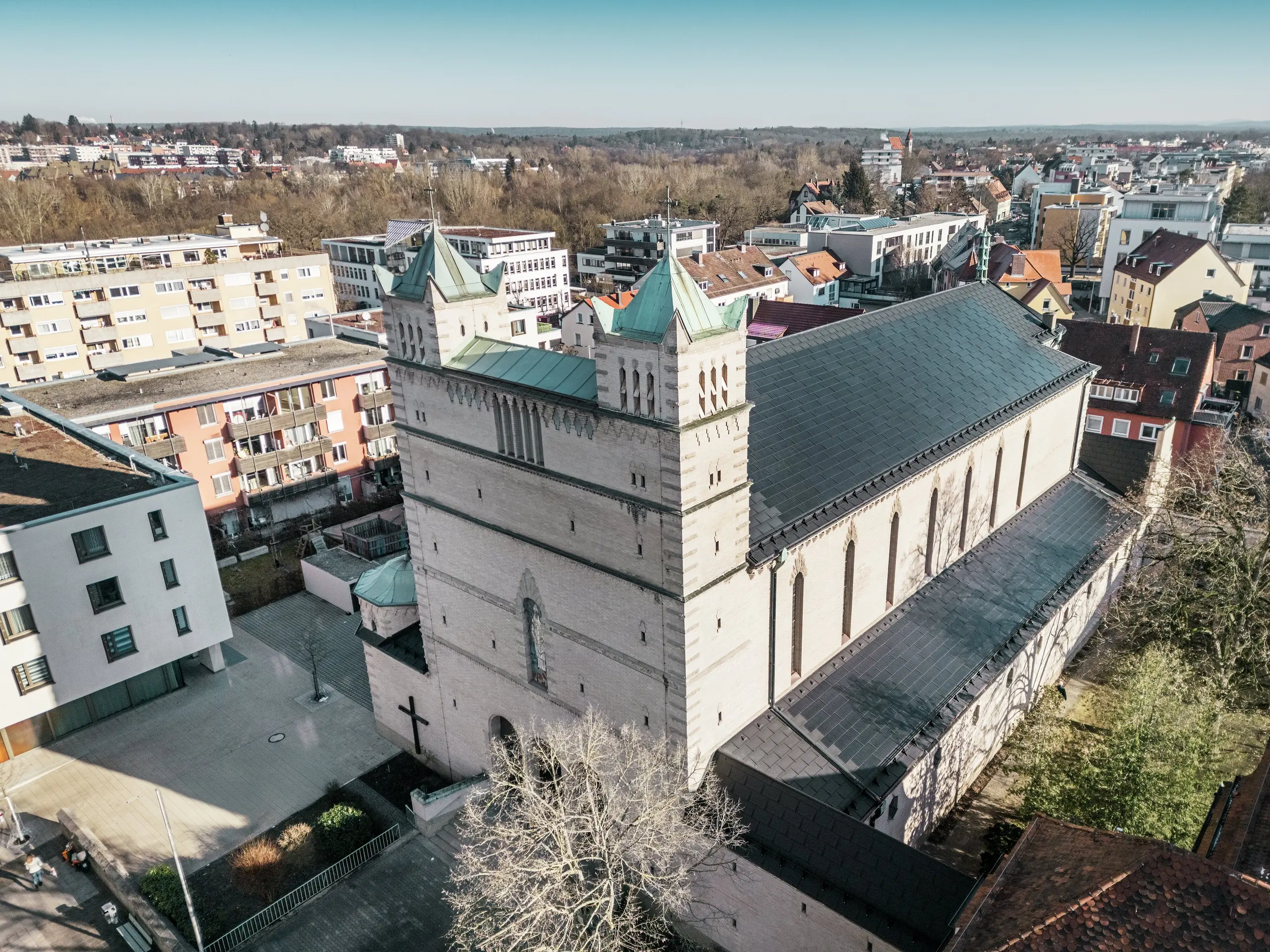 Sprednja stran cerkve sv. Karla Borromeja v Nürnbergu: slika prikazuje impresivno streho te cerkve, ki je zaščitena kot spomenik in pokrita s črnimi solarnimi strešnimi ploščami PREFA. Solarni moduli se harmonično vključujejo v zgodovinsko arhitekturo cerkve in hkrati predstavljajo sodobno rešitev za trajnostno pridobivanje energije. V ozadju so vidne sodobne stavbe in zeleni hribi Nürnberga, kar ponazarja uspešen prehod med tradicionalno cerkveno arhitekturo in sodobno oskrbo z energijo. Aluminijasto streho cerkve dopolnjuje elegantna PREFA strešna plošča  R.16.