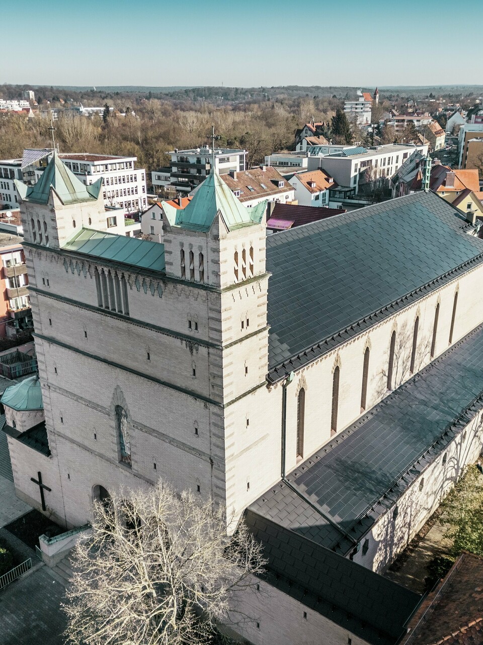 Vorderseite der St. Karl Borromäus Kirche in Nürnberg: Das Bild zeigt das beeindruckende Dach der denkmalgeschützten Kirche, das mit schwarzen PREFA-Solardachplatten gedeckt ist. Die Solarmodule fügen sich harmonisch in die historische Architektur der Kirche ein und stellen gleichzeitig eine moderne Lösung zur nachhaltigen Energiegewinnung dar. Im Hintergrund sind moderne Gebäude und die grünen Hügel Nürnbergs zu sehen, was den gelungenen Übergang zwischen traditioneller Kirchenarchitektur und zeitgemäßer Energieversorgung verdeutlicht. Das Aluminiumdach der Kirche wird durch die stilvolle PREFA Dachplatte R.16 vervollständigt.