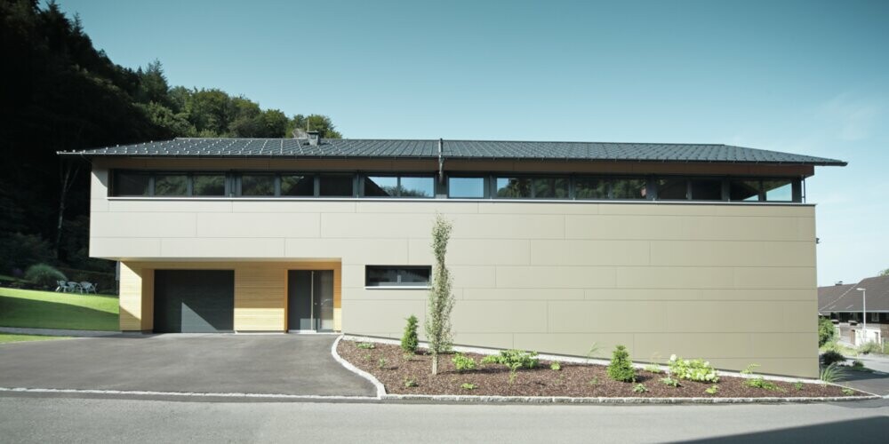 Dolga hiša s strešno ploščo PREFA v antracitni kritini P.10 v kombinaciji s kompozitno ploščo PREFA v bronasti barvi na fasadi; sodoben slog gradnje v kombinaciji z ozkimi okni in leseno fasado.