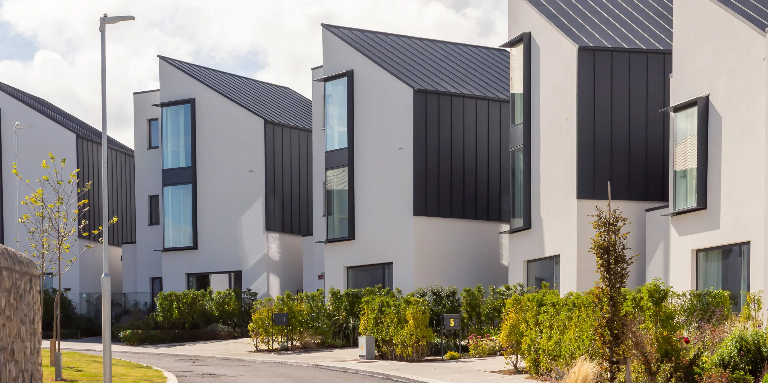 Moderne Reihenhausanlage Watson Place in der Nähe von Dublin mit klaren, asymmetrischen Dachformen und dunkler PREFALZ Verkleidung in P.10 Anthrazit. Die großformatigen Fenster und die markanten Aluminiumflächen betonen die dynamische Architektur. Die langlebigen PREFA Produkte bieten zuverlässigen Witterungsschutz und verleihen der Wohnanlage eine elegante, hochwertige Erscheinung.