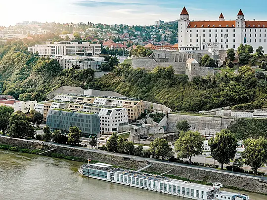 Pogled na četrt Vydrica v Bratislavi s pogledom na grad in Donavo: raznolika arhitektura navdušuje z zelenimi strešnimi površinami, strmimi dvokapnimi strehami in natančno vgrajenimi strešnimi okni, ki so vizualne posebnosti. PREFA aluminijaste strešne skodle v barvah prefa bela, peščeno rjava in bronasta povezujejo stavbe v harmonično celoto in se vklapljajo v zgodovinsko okolje. Trajne, lahke in izjemno vremensko odporne rešitve PREFA omogočajo kompleksne strešne geometrije in zagotavljajo trajnostno, vzdrževanju prijazno ovojnico stavbe z visoko oblikovno kakovostjo.