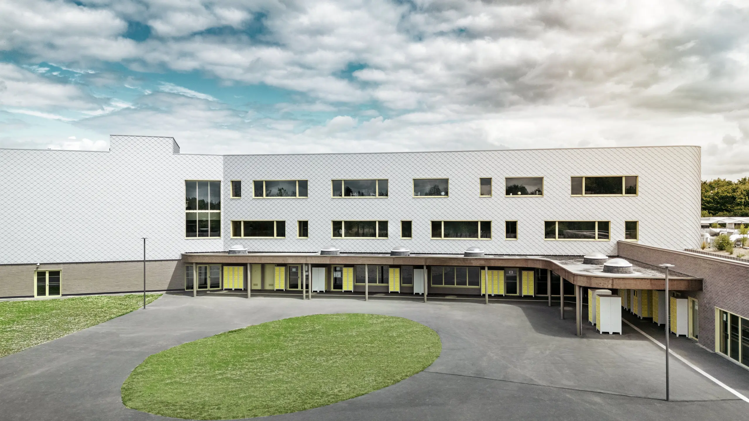 Zadnja stran šole Collège Léontine Dolivet z velikim šolskim dvoriščem in jasno arhitekturo. Fasada je sestavljena iz kovinsko srebrnih PREFA fasadnih rombov, ki poskrbijo za sodoben, eleganten videz. Več vrst oken in rumene vrata popestrijo strukturo, asfaltirano dvorišče s centralnim zelenim pasom pa ponuja veliko prostora za odmore. Trajna aluminijasta fasada združuje lahkotnost, odpornost proti vremenskim vplivom in dizajn – funkcionalna in estetsko privlačna rešitev v Cesson-Sévigné blizu Rennesa.