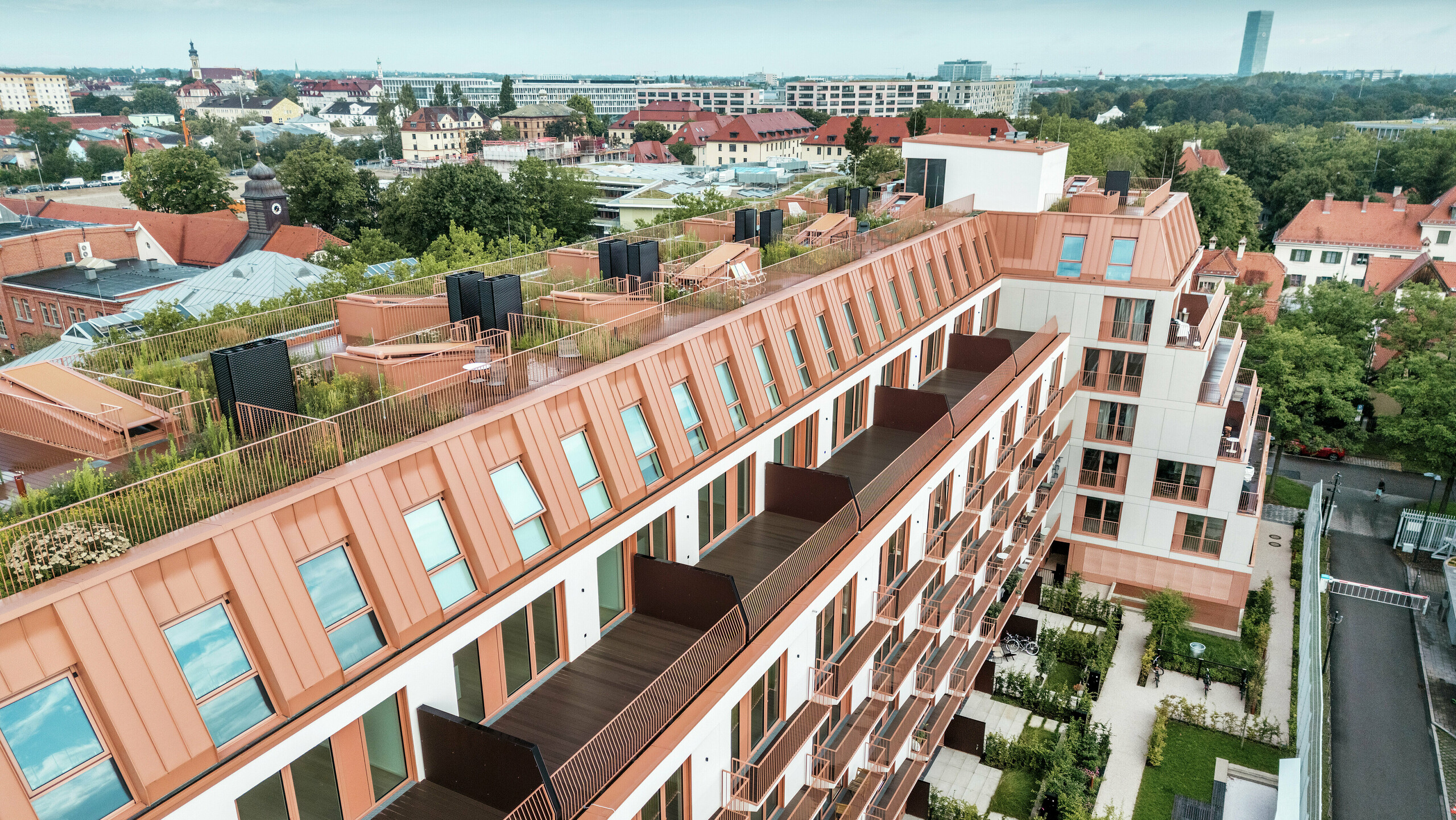 Pogled iz zraka na stanovanjski projekt Van B v Münchnu s presenetljivo aluminijasto streho PREFALZ v posebni barvi IGP Orange HDP podjetja PREFA. Prostorni balkoni, zelene strešne terase in natančna zasnova fasade poudarjajo premišljen arhitekturni koncept. Idealno za trajnostni urbani razvoj in urbano bivanje z visoko kakovostjo življenja in estetsko privlačnostjo.
