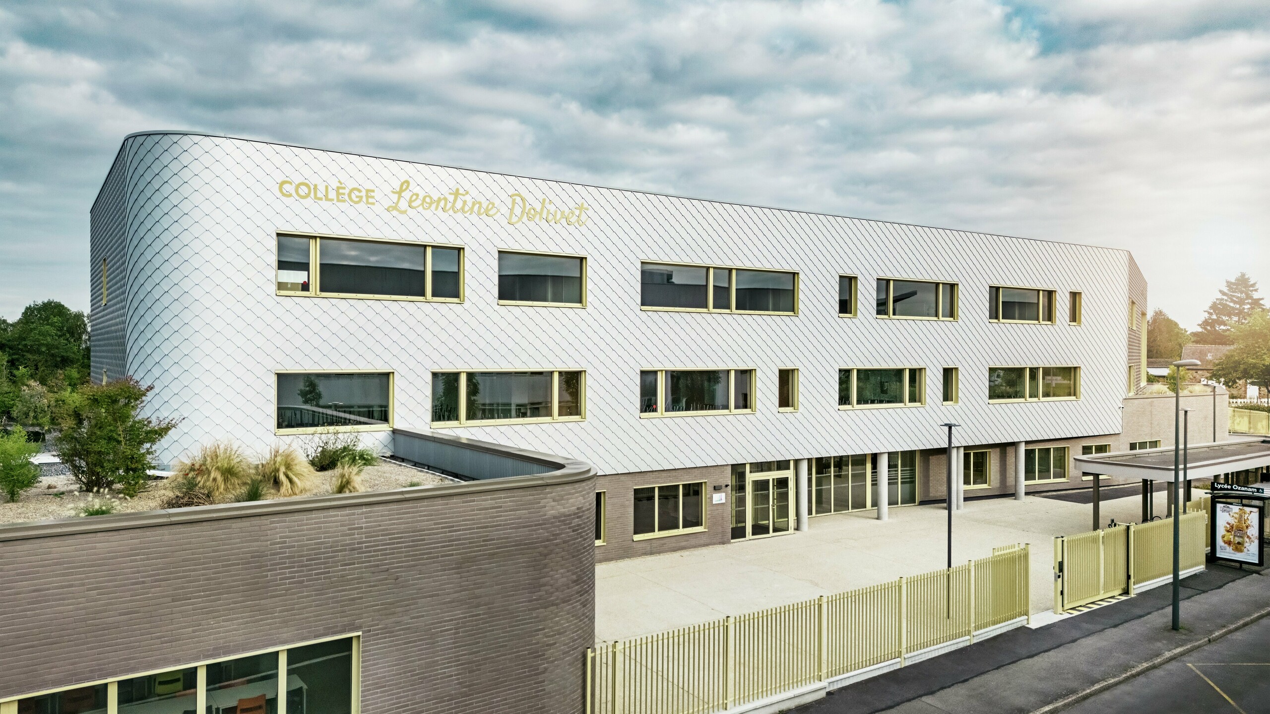 Sprednji pogled na Collège Léontine Dolivet v Cesson-Sévigné z moderno aluminijasto fasado iz PREFA fasadnih rombov v kovinsko srebrni barvi. Spreminjajoča se površina daje stavbi lahkotnost, medtem ko jasne linije in velika okna zagotavljajo preglednost. Rumeni okenski okvirji dajejo živahne poudarke. V ospredju so vidni svetel predprostor, ograja in sosednja cesta. Trajna fasadna rešitev PREFA združuje dizajn, vremensko odpornost in trajnost – arhitekturni magnet z elegantnim oblikovanjem.
