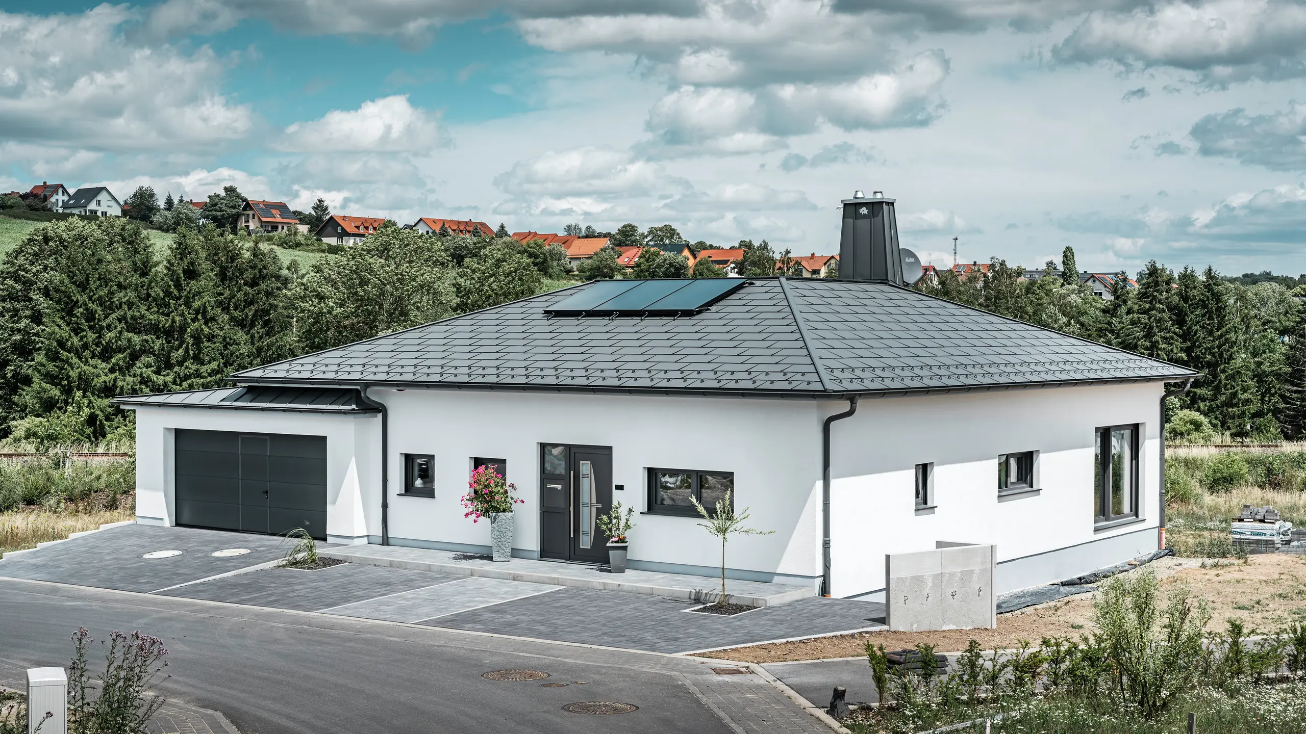 Elegantna enodružinska hiša v Ilmenauu v Turingiji z visokokakovostno PREFA streho v barvi P.10 temno siva, opremljeno z robustno strešno ploščo R.16. Garaža se po barvi in slogu harmonično ujema z uporabo PREFALZ. Pogled na posestvo pritegne funkcionalno oblikovano atrij in dimnik, okrašen s PREFA bikom, ki poudarja sodoben dizajn in inovativno strešno tehnologijo PREFA. Sončni kolektorji se brezhibno vključujejo v trajnostni koncept hiše. Prefinjen celoten koncept bungalova dopolnjuje PREFA sistem za odvodnjavanje strehe, ki je prav tako v temno sivi barvi.