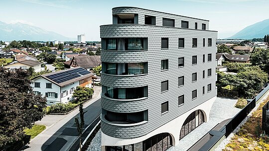 Letalski posnetek stavbe Bernerhaus v Buchsu z značilno trikotno obliko in nežno zaobljenimi balkoni. Fasada iz PREFA fasadnih skodel v barvi P.10 svetlo siva obdaja stavbo kot luskasta aluminijasta koža in poskrbi za živahno igro svetlobe in sence. Nad masivnim betonskim podstavkom z elegantnimi arkadami deluje lahka, vremensko odporna PREFA aluminijasta fasada še posebej natančna, trajna in enostavna za vzdrževanje – sodoben magnet za poglede sredi alpskega mestnega okolja.