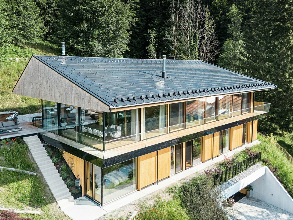 Pogled s ptičje perspektive na sodobno enodružinsko hišo v kraju Egg v Vorarlbergu s solarnimi strešnimi ploščami PREFA in strešnimi ploščami R.16 v črni barvi P.10. Aluminijasta strešna konstrukcija zagotavlja trajnostno pridobivanje energije in zagotavlja trdno zaščito pred vremenskimi vplivi. Zaščita pred snegom preprečuje zdrse snežnih mas na strehi v zimskih mesecih. Velika steklena fasada in lesena obloga zaokrožata privlačno zasnovo, v ozadju pa je vidna terasa s kakovostnim vrtnim pohištvom.