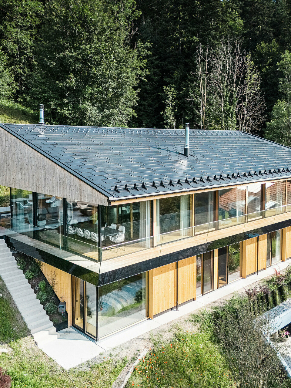 Pogled s ptičje perspektive na sodobno enodružinsko hišo v kraju Egg v Vorarlbergu s solarnimi strešnimi ploščami PREFA in strešnimi ploščami R.16 v črni barvi P.10. Aluminijasta strešna konstrukcija zagotavlja trajnostno pridobivanje energije in zagotavlja trdno zaščito pred vremenskimi vplivi. Zaščita pred snegom preprečuje zdrse snežnih mas na strehi v zimskih mesecih. Velika steklena fasada in lesena obloga zaokrožata privlačno zasnovo, v ozadju pa je vidna terasa s kakovostnim vrtnim pohištvom.