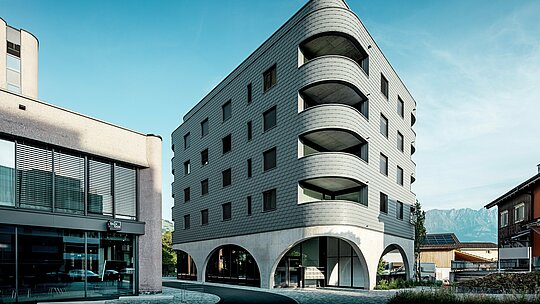 Ta sodobna stavba Bernerhaus v Buchsu navdušuje s svojo izrazito zaobljeno, trikotno obliko stavbe in neprekinjeno fasado iz PREFA fasadnih skodel v barvi P.10 svetlo siva. Luskasta aluminijasta površina poudarja mehke robove in globoke zaobljene balkone, medtem ko masivni betonski podstavek z elegantnimi arkadami ustvarja zanimiv kontrast. Pred alpskim ozadjem sredi majhne nakupovalne ulice postane lahka, vremensko odporna in trajna PREFA aluminijasta fasada arhitekturni magnet za poglede: natančno je izdelana, zahteva malo vzdrževanja in je idealna za zahtevne zaobljene oblike.