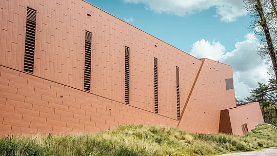Značilen stranski pogled na črpališče pitne vode WML v Lottumu z jasno razčlenjenim, podolgovatim objektom sredi naravne travniške pokrajine. Aluminijasta fasada PREFA Siding.X v topli rjavi barvi je strukturirana z ritmično razporejenimi, vitkimi lamelami, ki ustvarjajo živahno igro svetlobe in sence. Fino fasetirana površina deluje, odvisno od perspektive, dinamično in kakovostno. Lahki, trajni in v celoti reciklirani PREFA aluminijasti paneli zagotavljajo visoko odpornost proti vremenskim vplivom, barvno obstojnost ter trajnostno, vzdrževanju prijazno ovojnico stavbe.