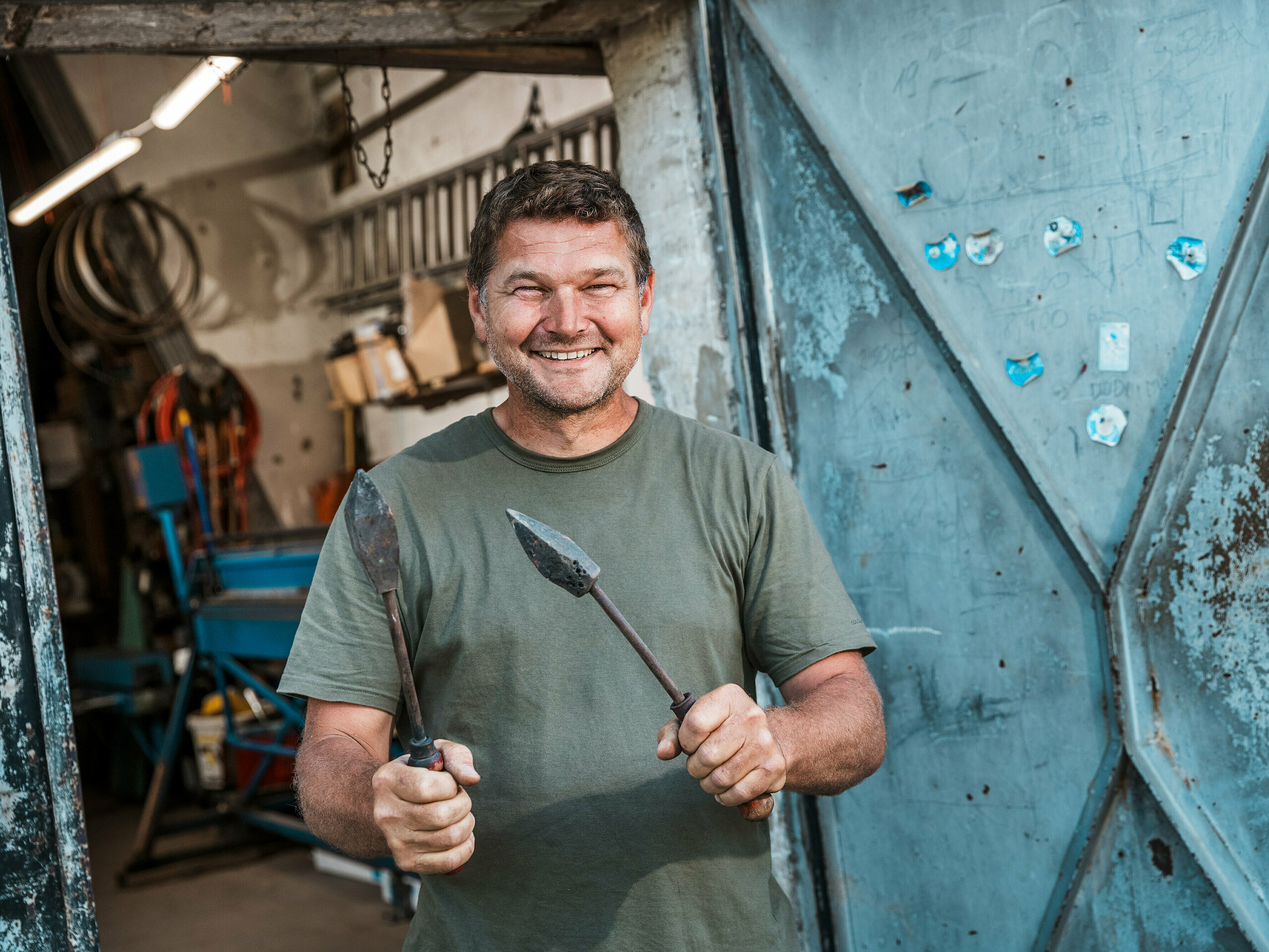 Krovec Richárd Tóth v svoji delavnici. Ta priznani obrtnik drži v rokah tradicionalno orodje in stoji pred odprtimi kovinskimi vrati. Prizor izžareva pristno obrtništvo in visoko natančnost – lastnosti, ki so bile ključnega pomena pri obdelavi PREFA aluminijastih izdelkov za večstanovanjsko stavbo v Budimpešti. Robustno okolje delavnice poudarja obrtniško strokovnost, s katero so bili izdelani fasadni elementi, atika in balkonske obloge iz PREFALZ v barvi P.10 antracit. Slika poudarja povezavo med izkušnjami, poznavanjem materialov in trajnimi rešitvami PREFA.