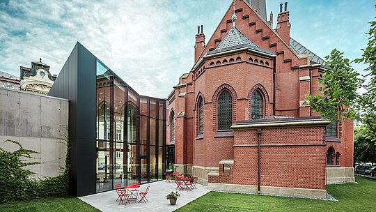 Slika prikazuje impresivno novo stavbo, ki povezuje zgodovinsko Rdečo cerkev z moderno raziskovalno knjižnico v Olomoucu. Perspektiva prikazuje zadnjo stran stavbe z vabljivo teraso. Elegantna fasada iz črnih PREFABOND aluminijastih kompozitnih plošč daje izrazit, sodoben poudarek in je v stilskem kontrastu z rdečo opeko cerkve. Visoka natančnost montaže in uporaba trajnih materialov, kot je aluminij, zagotavljajo harmonično povezavo tradicije in inovativnosti. Te napredne plošče niso le estetsko privlačne, ampak tudi izjemno funkcionalne in trajne, kar projekt spreminja v mojstrovino moderne arhitekture.