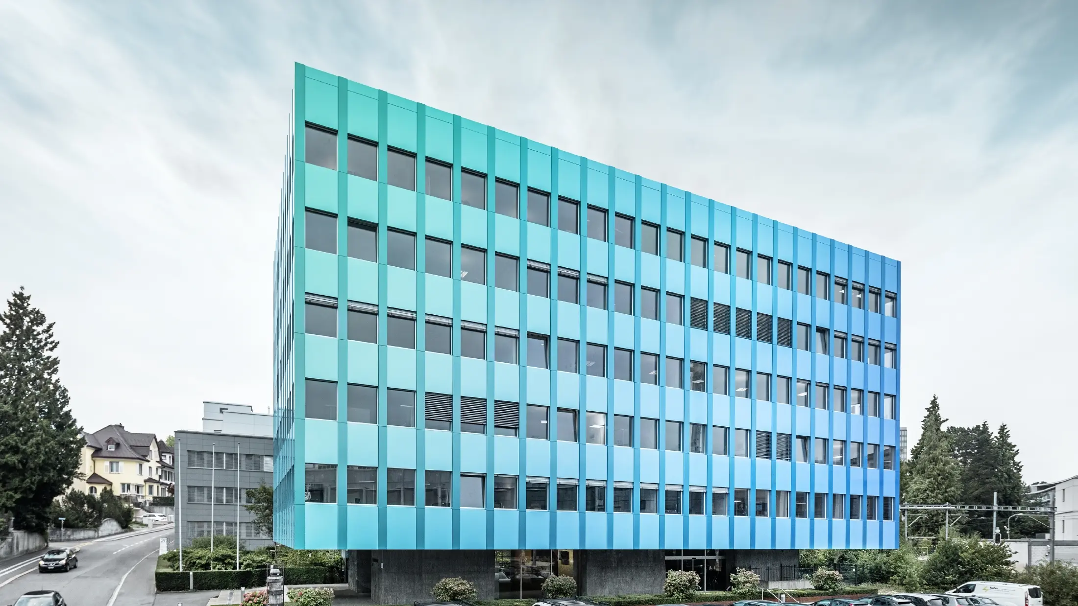 Stranski pogled na Wielandovo hišo v Aarau; fasada je obložena s PREFABOND aluminijasto kompozitno ploščo proizvajalca PREFA, ki zaradi posebnega postopka premazovanja mavrično modro-zeleno.