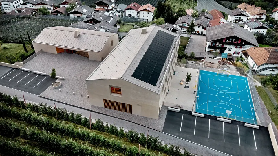 Pogled iz zraka na izobraževalni center Riffian na Južnem Tirolskem z vrtcem, osnovno šolo in športnim igriščem. Strešni rombi PREFA 44 × 44 v barvi P.10 bronasta tvorijo natančno izvedeno in trajno aluminijasto strešno kritino, ki se harmonično vključuje v alpsko pokrajino. Sodobno arhitekturo odlikujejo odpornost proti vremenskim vplivom in čiste oblikovne linije, premišljene do najmanjše podrobnosti.