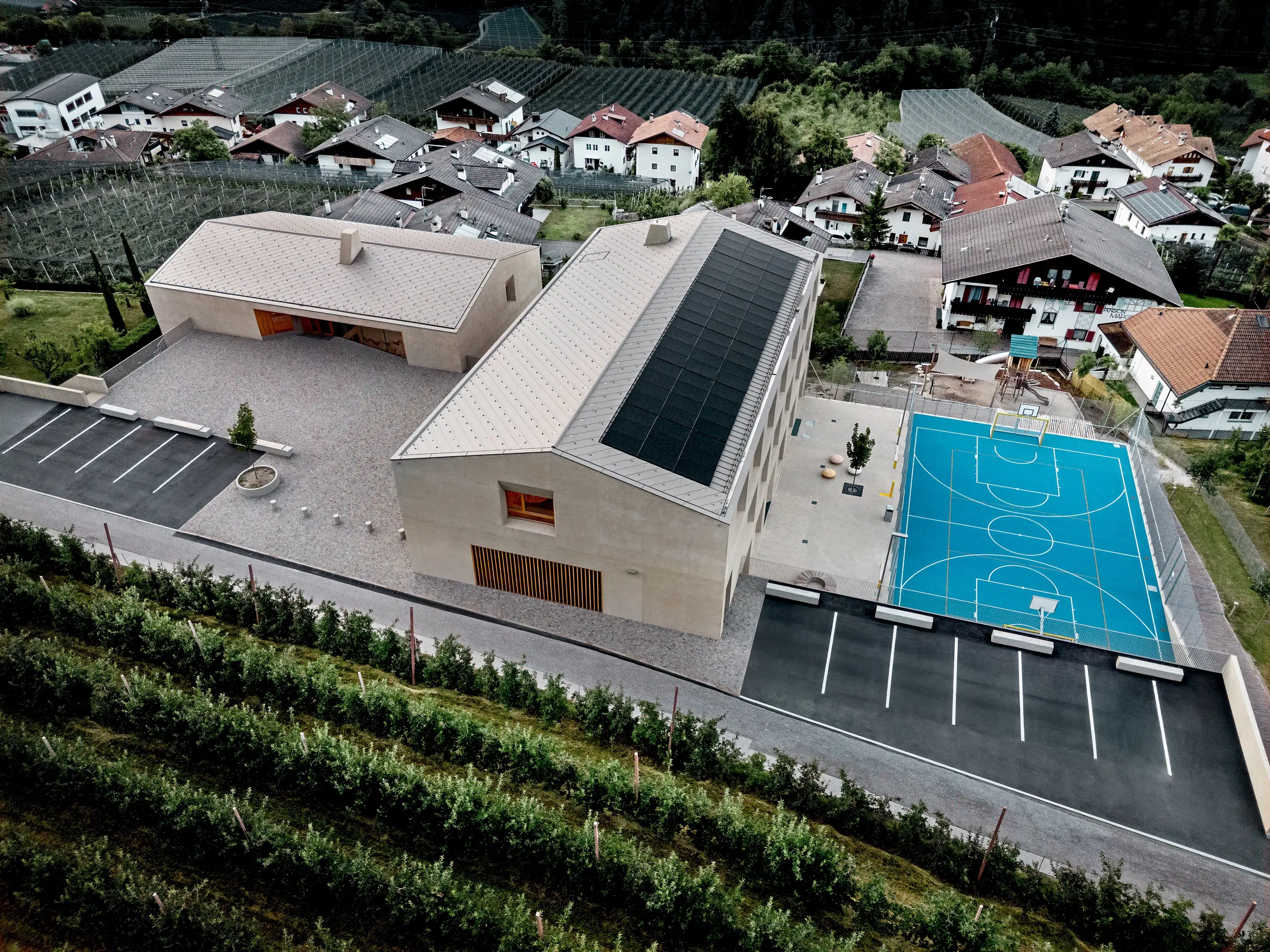 Pogled iz zraka na izobraževalni center Riffian na Južnem Tirolskem z vrtcem, osnovno šolo in športnim igriščem. Strešni rombi PREFA 44 × 44 v barvi P.10 bronasta tvorijo natančno izvedeno in trajno aluminijasto strešno kritino, ki se harmonično vključuje v alpsko pokrajino. Sodobno arhitekturo odlikujejo odpornost proti vremenskim vplivom in čiste oblikovne linije, premišljene do najmanjše podrobnosti.
