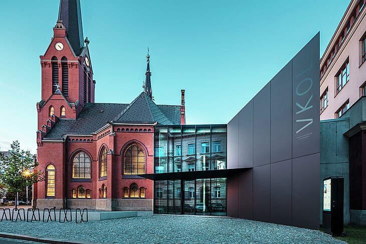 Slika prikazuje impresivno prepletanje zgodovinske Rdeče cerkve in moderne nove stavbe v Olmützu. Cerkev s svojimi značilnimi gotskimi okni in izrazitim stolpom tvori zgodovinsko hrbtenico kompleksa, medtem ko moderni prizidek s svojo elegantno črno fasado iz aluminijastih kompozitnih plošč PREFABOND predstavlja sodoben arhitekturni odgovor. Velike steklene površine omogočajo pogled v notranjost nove stavbe, kjer okolica z odsevi v oknih odraža zanimivo interakcijo med starim in novim. Natančna obdelava plošč PREFABOND poudarja sodobno estetiko, medtem ko harmonična integracija v zgodovinsko okolje navdušuje tako funkcionalno kot vizualno. 