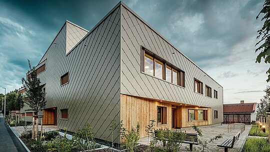 V prijetnem večernem vzdušju se hibridna stavba vrtca v mestu Alzenau razkriva v vsej svoji arhitekturni veličini. Izrazita fasada iz bronastih PREFA fasadnih rombov 44 × 44 daje stavbi eleganten, vremensko odporen ovoj z jasno strukturo. V pritličju vertikalne lesene obloge ustvarjajo naraven, topel kontrast z aluminijem in poudarjajo vhodni prostor ter zaščitene bivalne cone. Minimalistična, moderna oblika se harmonično ujema z urejenim zunanjim prostorom, kjer klopi, stojala za kolesa in mlada drevesa vabijo k počitku. Slika nazorno prikazuje, kako trajni izdelki PREFA prispevajo k trajnostni, funkcionalni in hkrati vabljivi izobraževalni stavbi.