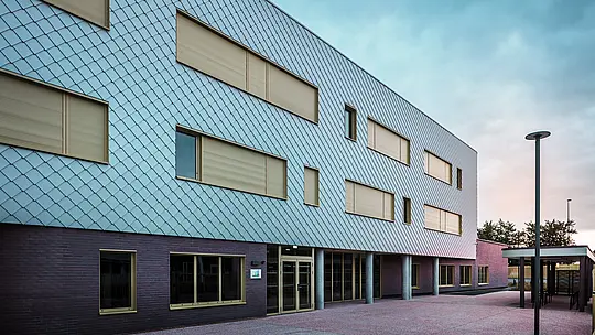 Fotografija prikazuje vhodno stran šole Collège Léontine Dolivet z moderno aluminijasto fasado iz PREFA fasadnih rombov v kovinsko srebrni barvi. Več pravokotnih oken z rumenimi okvirji in zaprtimi roletami strukturira pogled. V ospredju je tlakovano preddverje, obdano s pokritim prostorom. Fasada deluje elegantno, je odporna na vremenske vplive in trajna.