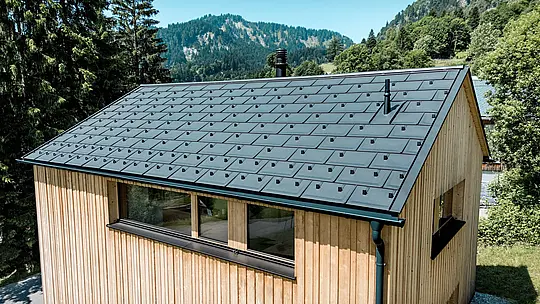 V svetlem gorskem poletju črna aluminijasta streha PREFA na hiši Immi na impresiven način stopa v središče pozornosti. Solarne strešne plošče, vgrajene tako, da so v isti ravnini s streho, skupaj s strešnimi ploščami R.16 v barvi P.10 črna tvorijo mirno, enotno strešno površino. Lahki, robustni aluminijasti sistem proizvaja sončno energijo in leseno hišo zanesljivo ščiti pred zahtevnimi alpskimi vremenskimi razmerami ter velikimi snežnimi obremenitvami.